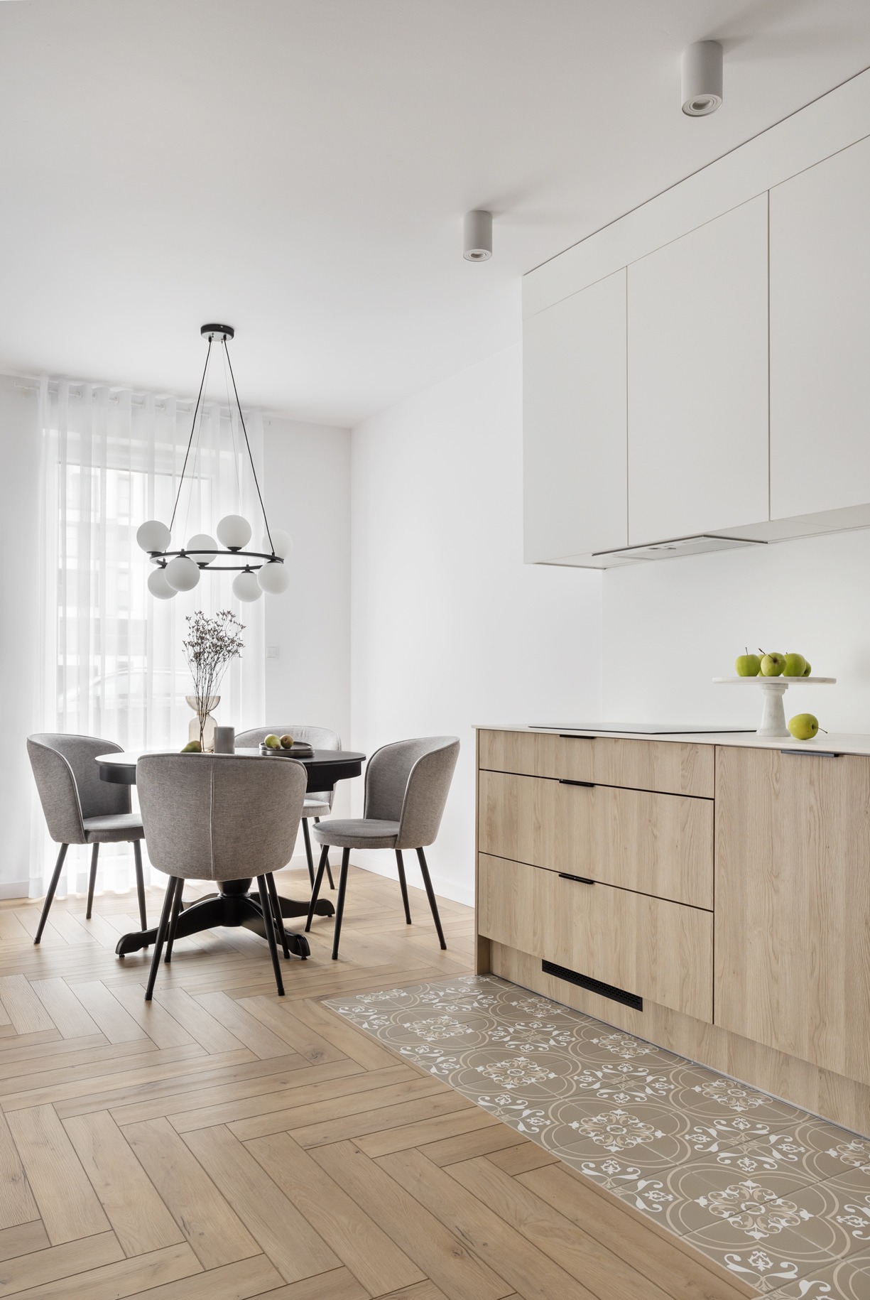 Jasna kuchnia z drewnianymi szafkami i czarnym stołem otoczonym szarymi krzesłami, parkiet w jodełkę i dekoracyjna mata pod szafkami.