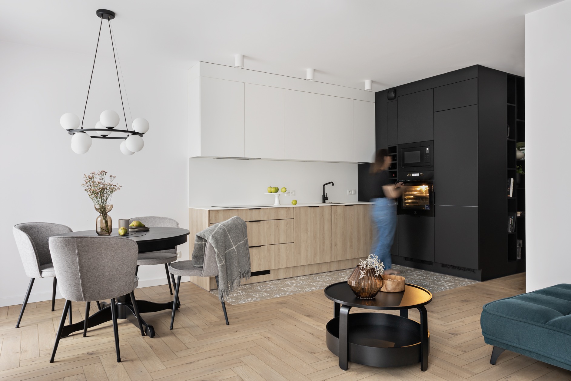 Jasna kuchnia z drewnianymi frontami szafek dolnych i białymi górnymi, czarna zabudowa z piekarnikiem, okrągły stół z szarymi krzesłami, parkiet w jodełkę, kobieta wyjmująca potrawę z piekarnika.