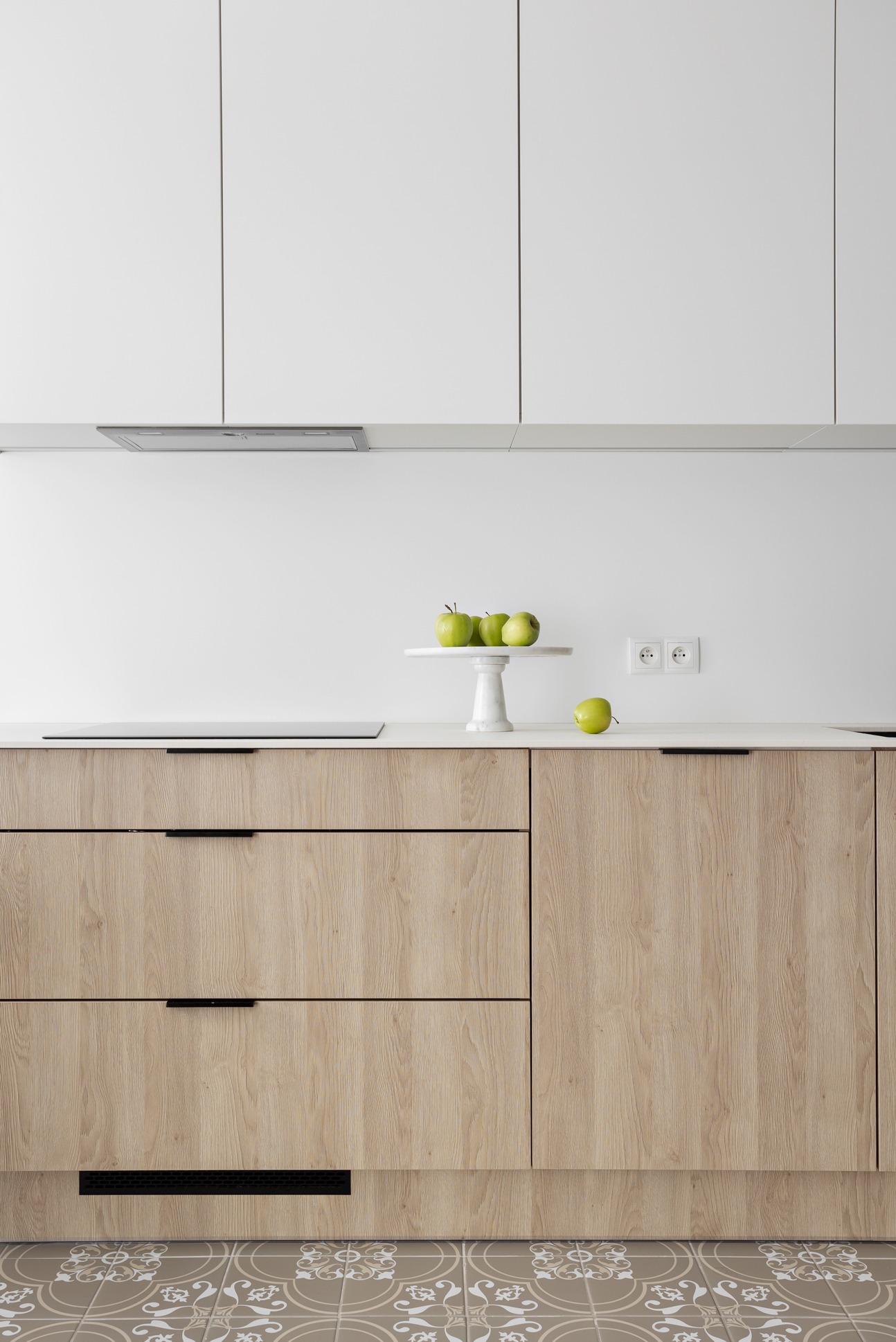 Minimalistyczna kuchnia: białe szafki górne, drewnopodobne dolne z czarnymi uchwytami, marmurowy cokół z zielonymi jabłkami, wzorzysta podłoga i wbudowany okap