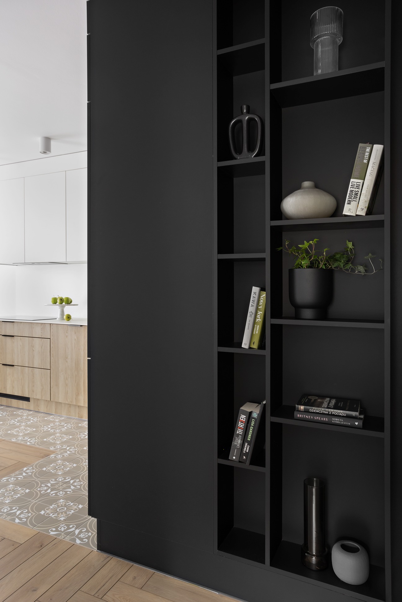 Częściowy widok nowoczesnego mieszkania: czarny regał z dekoracjami (wazony, książki, roślina), fragment kuchni z drewnianymi frontami i blatem, podłoga z płytek wzorzystych i parkietu.