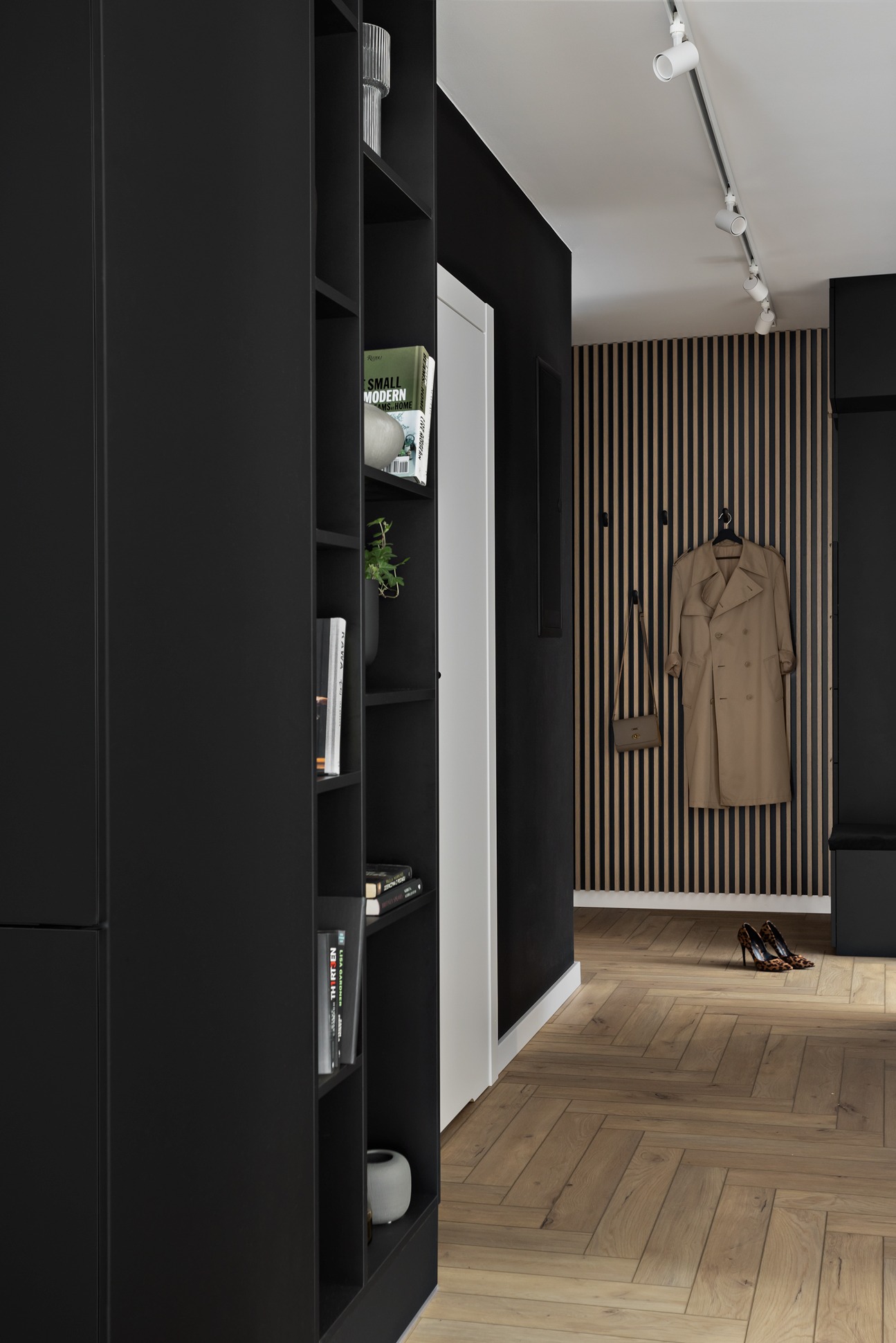Wnętrze przedpokoju z czarnymi meblami, drewnianą ścianą z wieszakami, beżowym płaszczem, torebką i damskimi butami w panterkę na podłodze z parkietu w jodełkę.