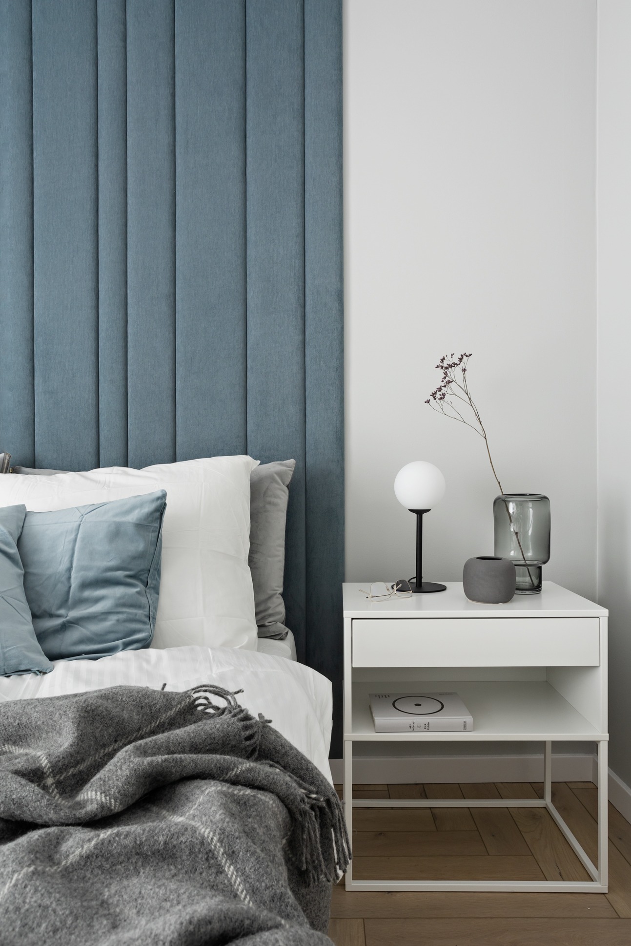 Sypialnia z niebieskim, tapicerowanym panelem zagłówkowym, białą pościelą, szarą narzutą w kratę i białą szafką nocną z lampką i dekoracjami.