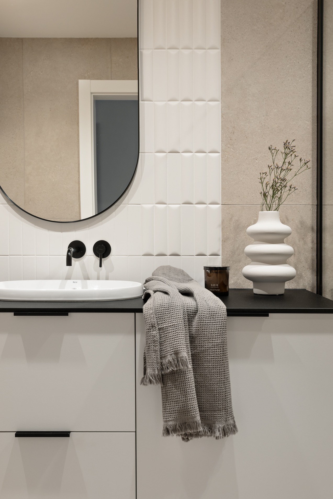 Nowoczesna łazienka w Gliwicach z owalnym lustrem w czarnej ramie, białą umywalką, czarną armaturą, teksturowanymi białymi płytkami i beżową ścianą, uzupełniona o wazon z suszonymi kwiatami i szary...