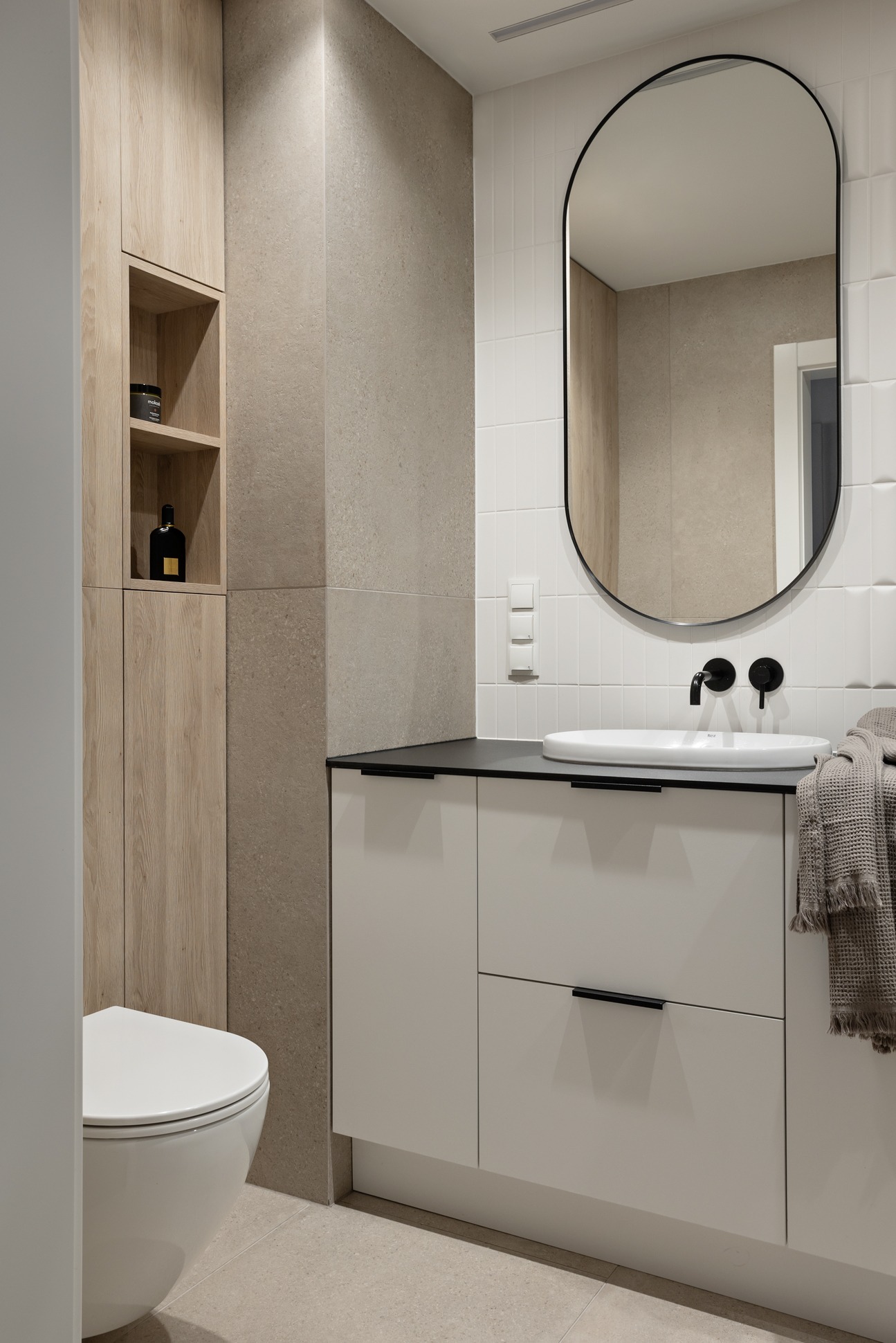 Nowoczesna łazienka z owalnym lustrem, białą umywalką nablatową z czarną armaturą i szafką pod umywalkową z szufladami, obok toaleta podwieszana i drewniana szafka z półkami.