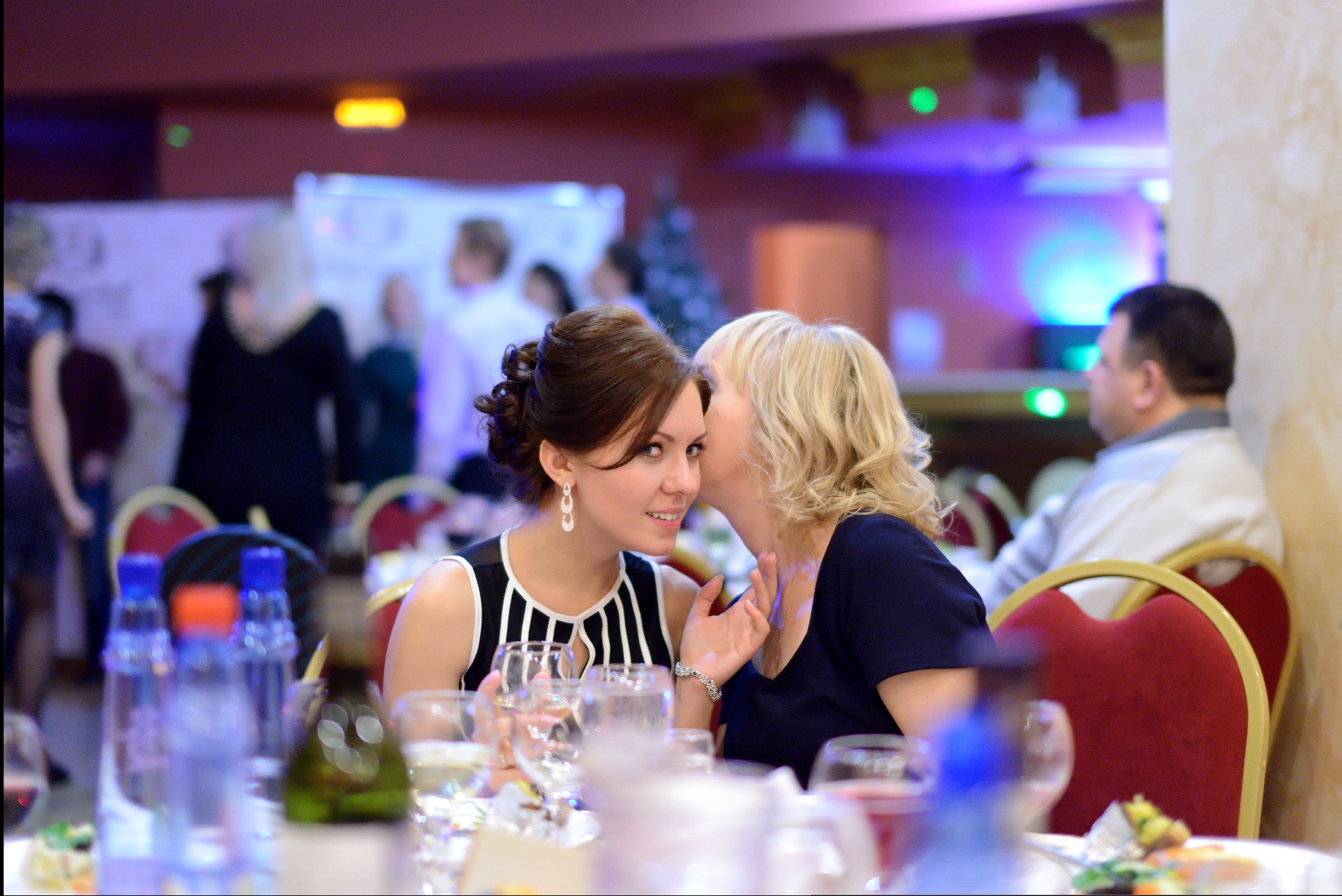 Dwie kobiety siedzą przy stole podczas weselnego przyjęcia; blondynka szepcze coś do ucha brunetce w sukience w biało-czarne paski, która uśmiecha się patrząc w obiektyw; na stole szklanki, butelki...