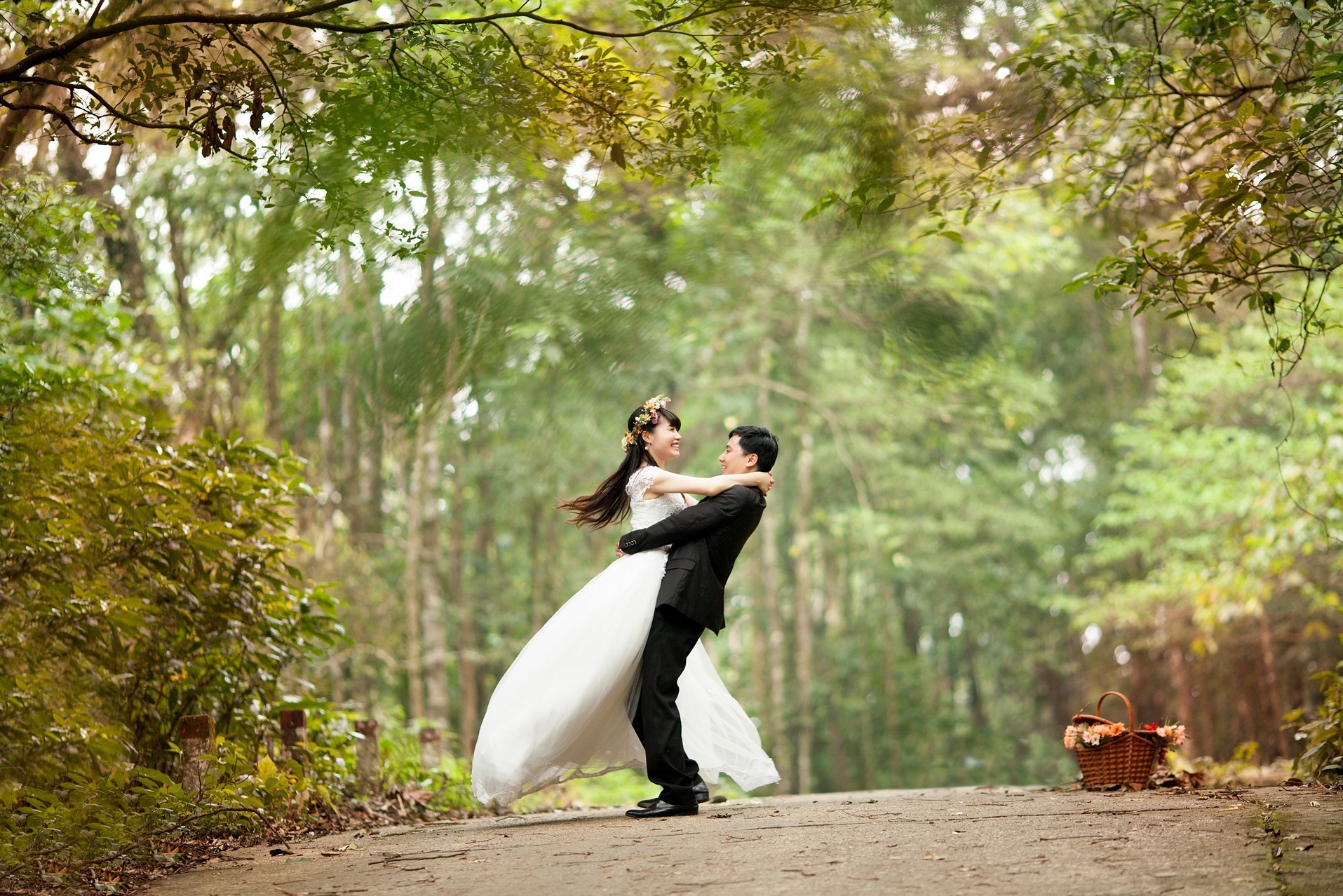 Uśmiechnięta panna młoda w białej sukni i wianku kwiatów na głowie, obejmowana przez pana młodego w czarnym garniturze na leśnej ścieżce, koszyk z kwiatami w tle.
