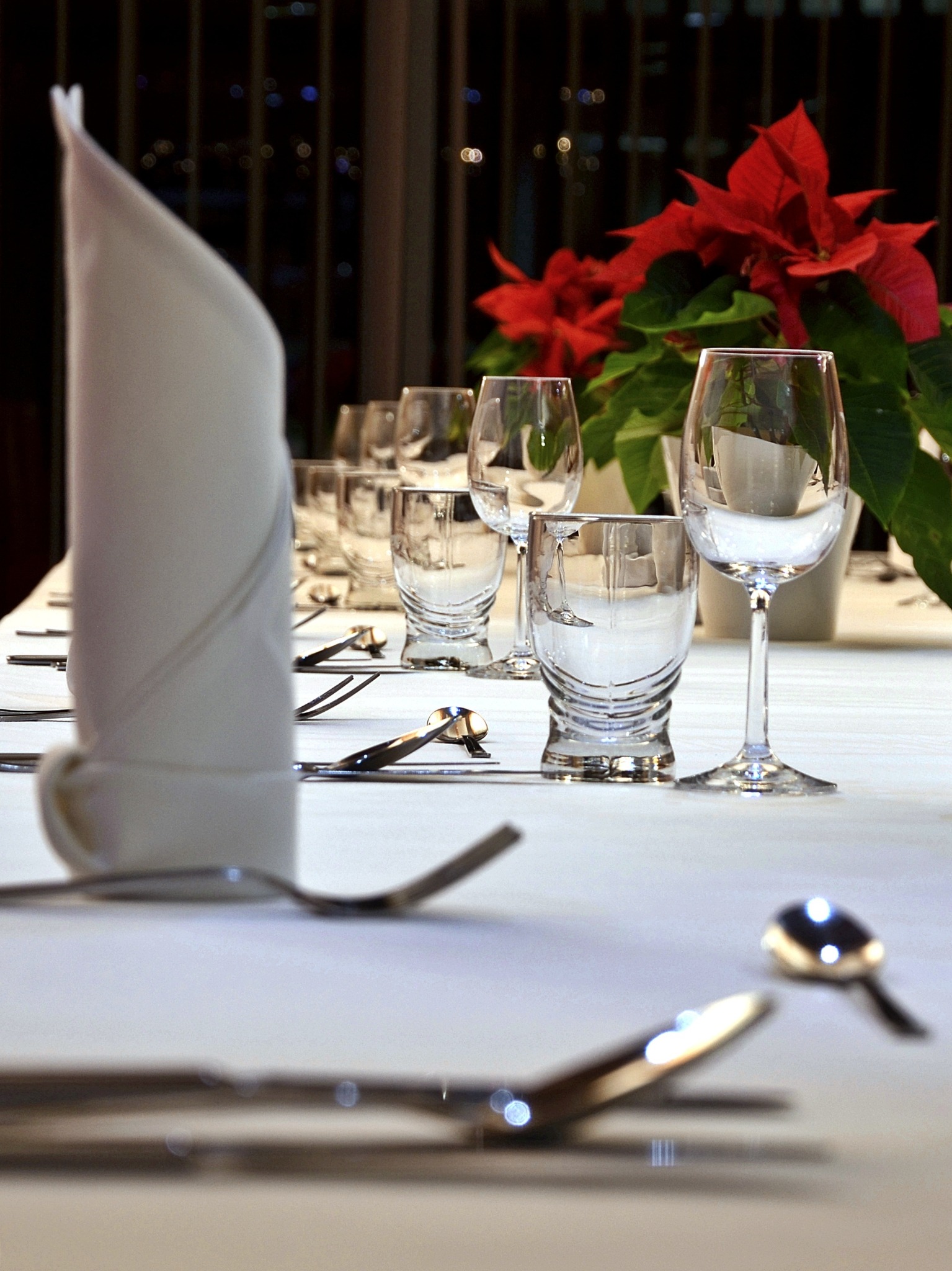Elegancko nakryty stół z białym obrusem, złożonymi serwetkami, szklankami, kieliszkami i sztućcami, udekorowany czerwonymi gwiazdami betlejemskimi, z rozmytym tłem miejskich świateł.