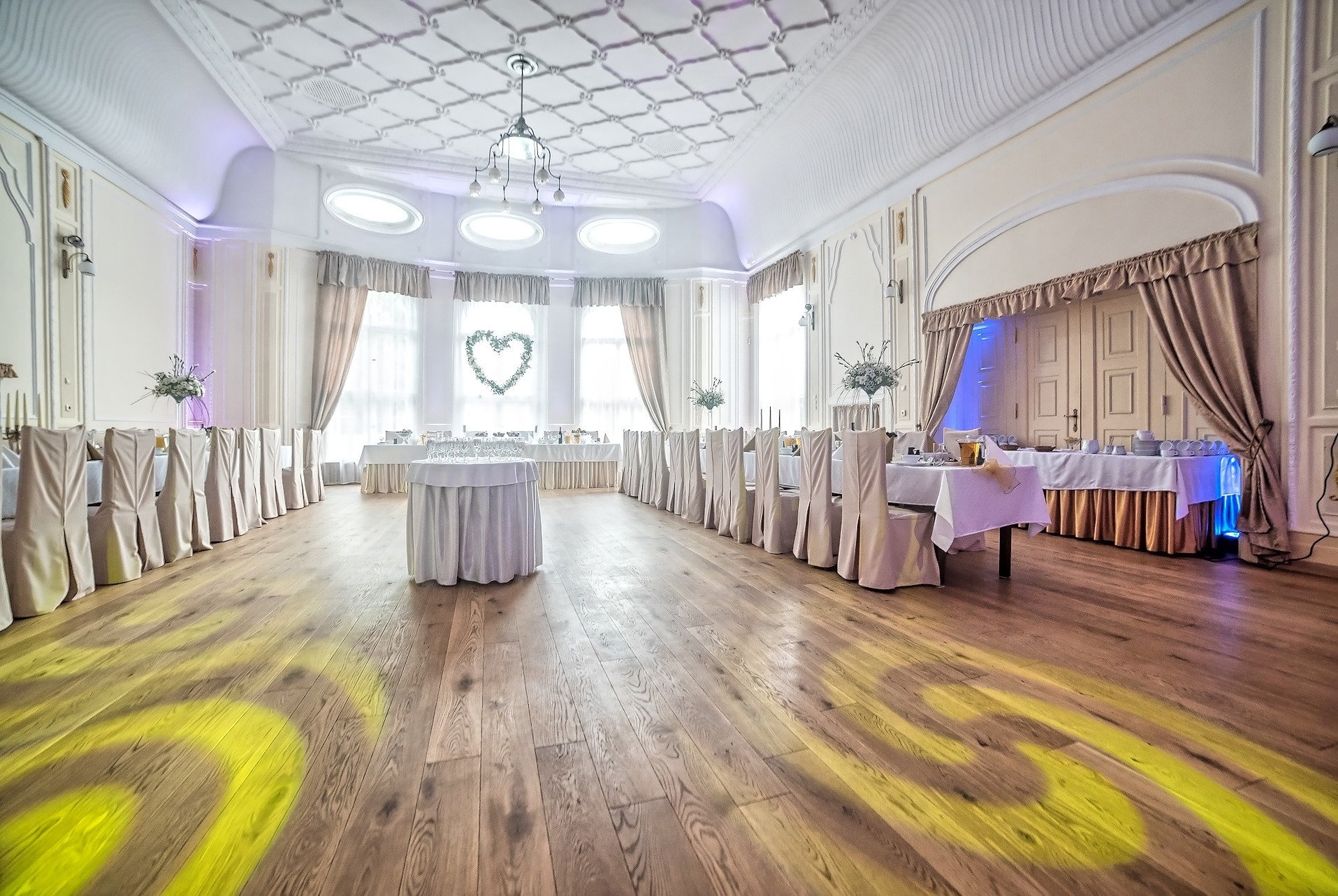 Uroczysta sala weselna z długimi stołami, białymi obrusami i krzesłami w pokrowcach, dekorowana kwiatami i sercem z roślin w oknie, oświetlona parkietowymi reflektorami.