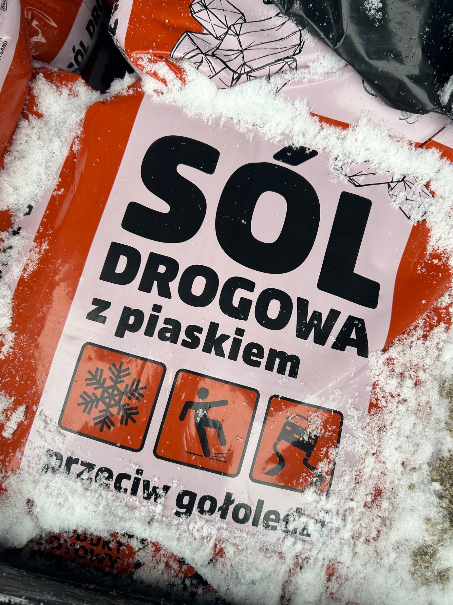 Worek z solą drogową z piaskiem, pokryty warstwą śniegu, widoczne symbole ostrzegawcze przed gołoledzią i utratą przyczepności.