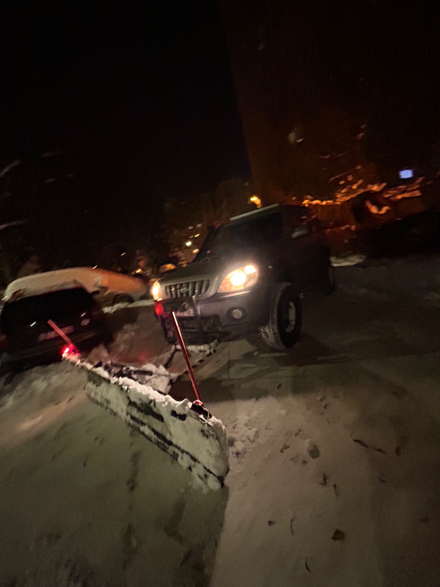 Ciemny samochód z pługiem odśnieżającym na zaśnieżonej drodze w nocy, z widocznymi światłami i innymi samochodami w tle.