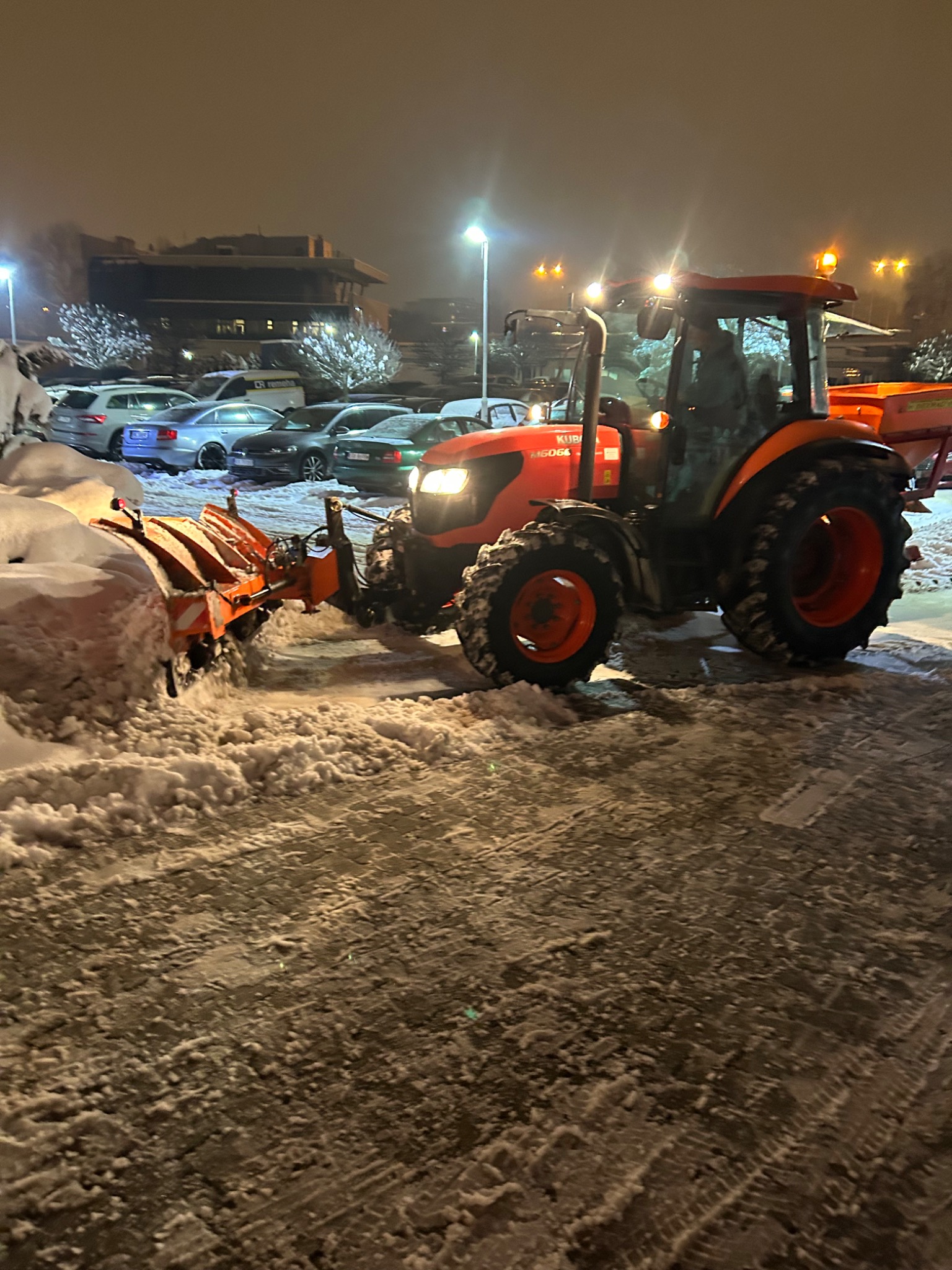 Pomarańczowy traktor Kubota z pługiem odśnieżającym usuwa śnieg z parkingu w nocy, w tle zaparkowane samochody i oświetlony budynek.