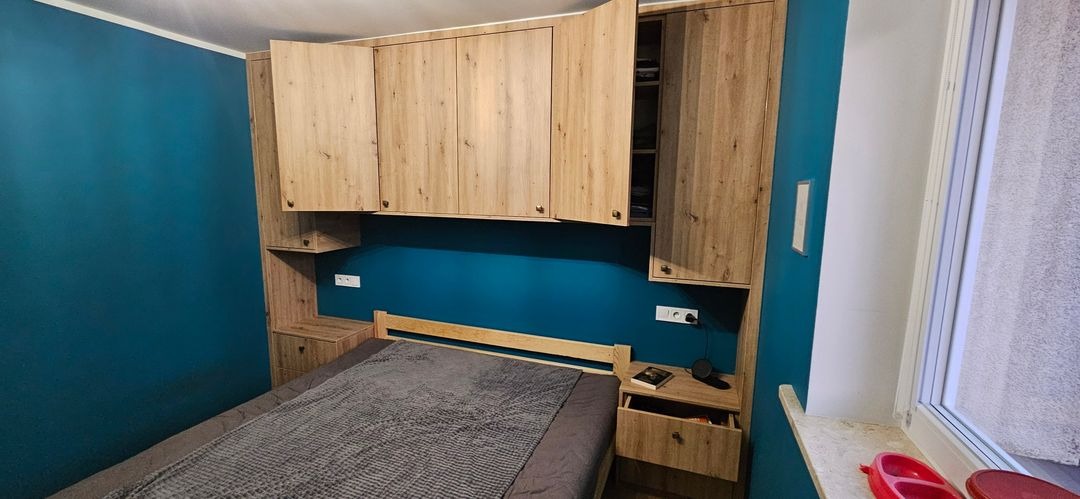 Zabudowa szaf nad łóżkiem w sypialni w Bielsku-Białej, z otwartą szafką ukazującą zawartość, drewnopodobny dekor frontów, turkusowa ściana w tle.