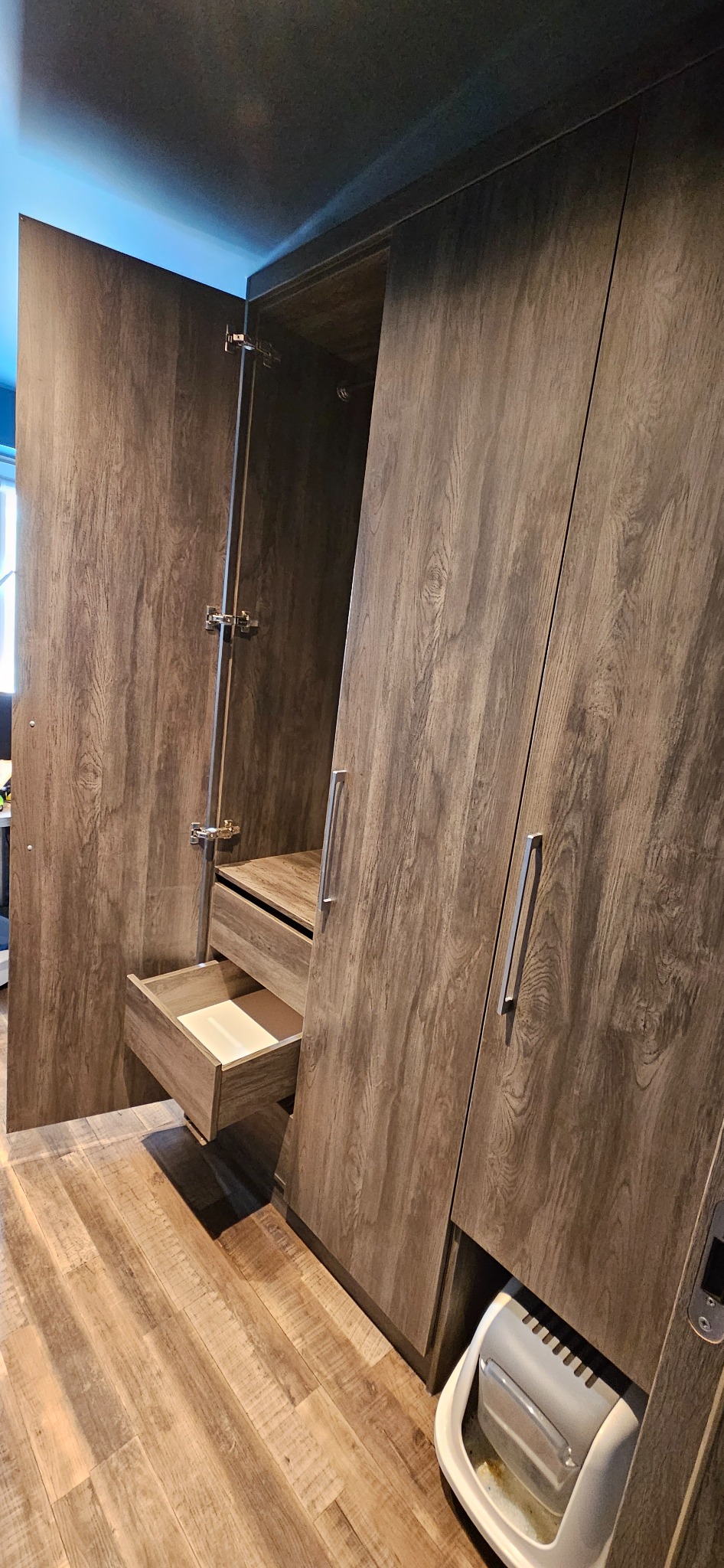 Drewniana szafa z otwartymi drzwiami i wysuniętą szufladą, na podłodze panele imitujące drewno, w dolnej części szafy widoczny pojemnik.