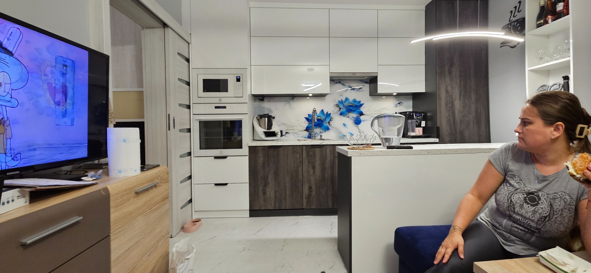 Nowoczesna kuchnia z białymi szafkami górnymi i ciemnymi szafkami dolnymi z motywem drewna, ozdobiona panelem szklanym z motywem niebieskich kwiatów; kobieta siedzi z hamburgerem na pierwszym...