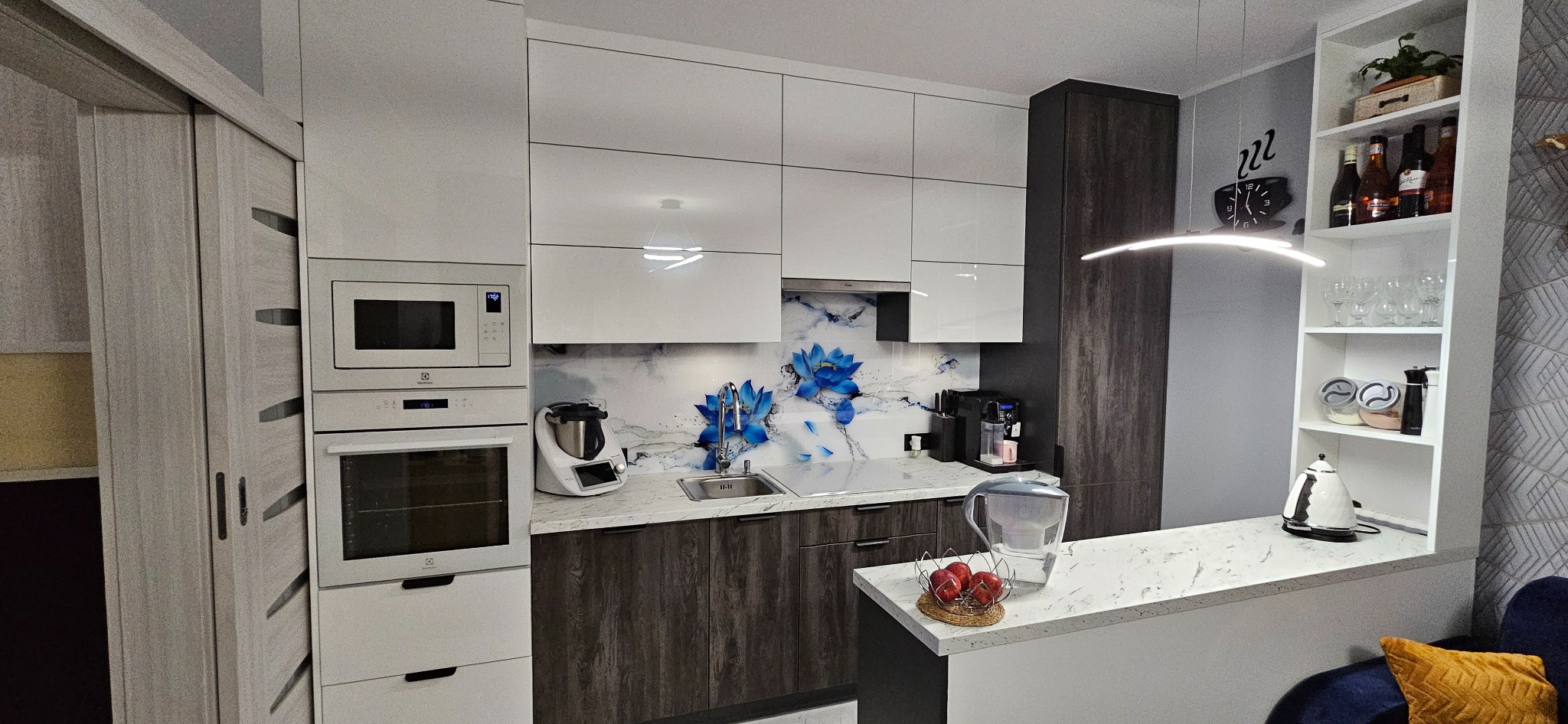 Nowoczesna kuchnia z białymi, błyszczącymi szafkami górnymi i ciemnymi, drewnopodobnymi szafkami dolnymi, zintegrowanym sprzętem AGD Electrolux i dekoracyjnym panelem nad blatem z motywem...