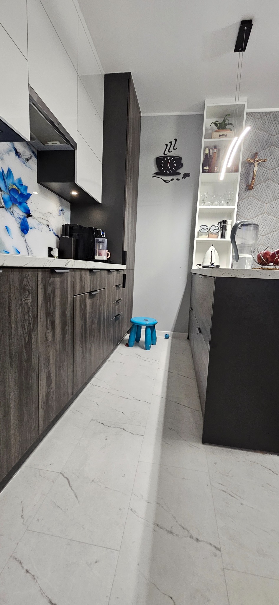 Nowoczesna kuchnia z szarymi szafkami z drewnianą teksturą, białymi szafkami wiszącymi i blatem z marmurowym wzorem; na ścianie zegar w kształcie filiżanki kawy, a na podłodze niebieski taboret.