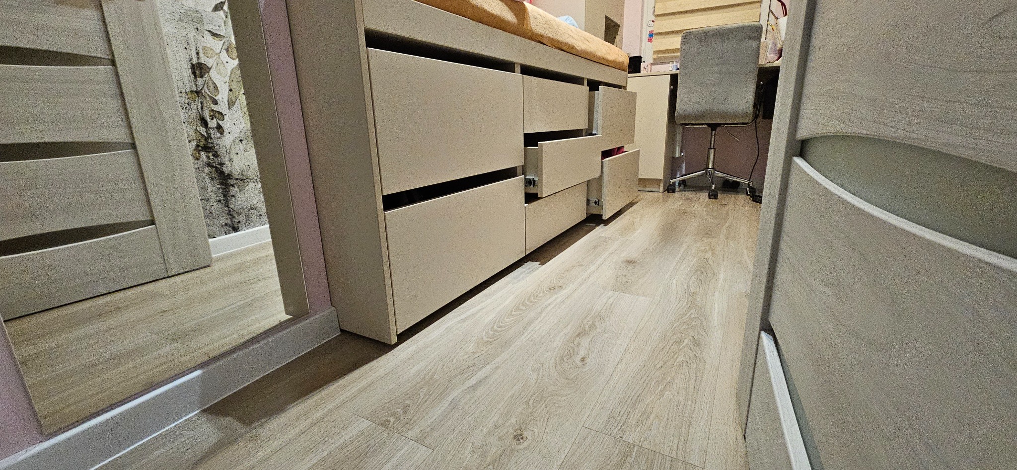 Beżowa komoda z szufladami, niektóre otwarte, na jasnej drewnianej podłodze, widoczna część biurka z krzesłem w tle, odbicie w lustrze na ścianie po lewej stronie.