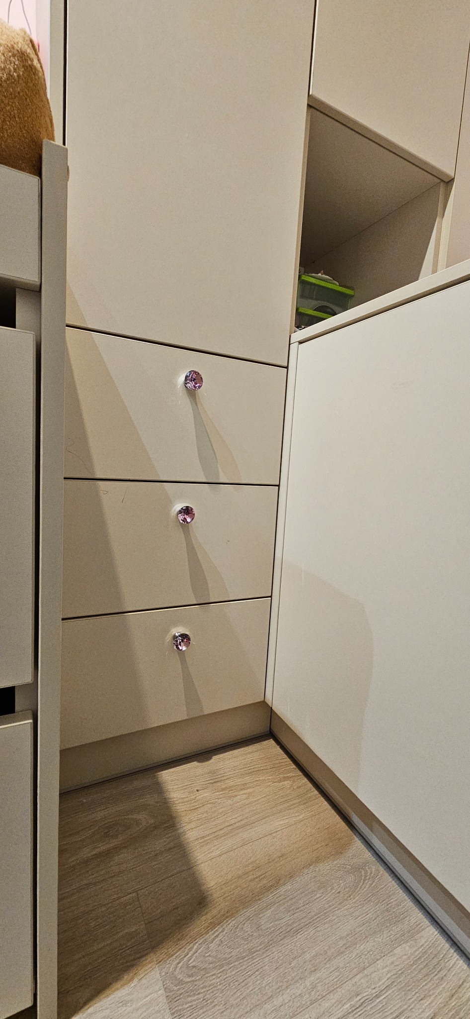 Fragment zabudowy meblowej z trzema szufladami, każda z ozdobnym, kryształowym uchwytem w kolorze różowym, widoczne również półki i podłoga imitująca drewno.