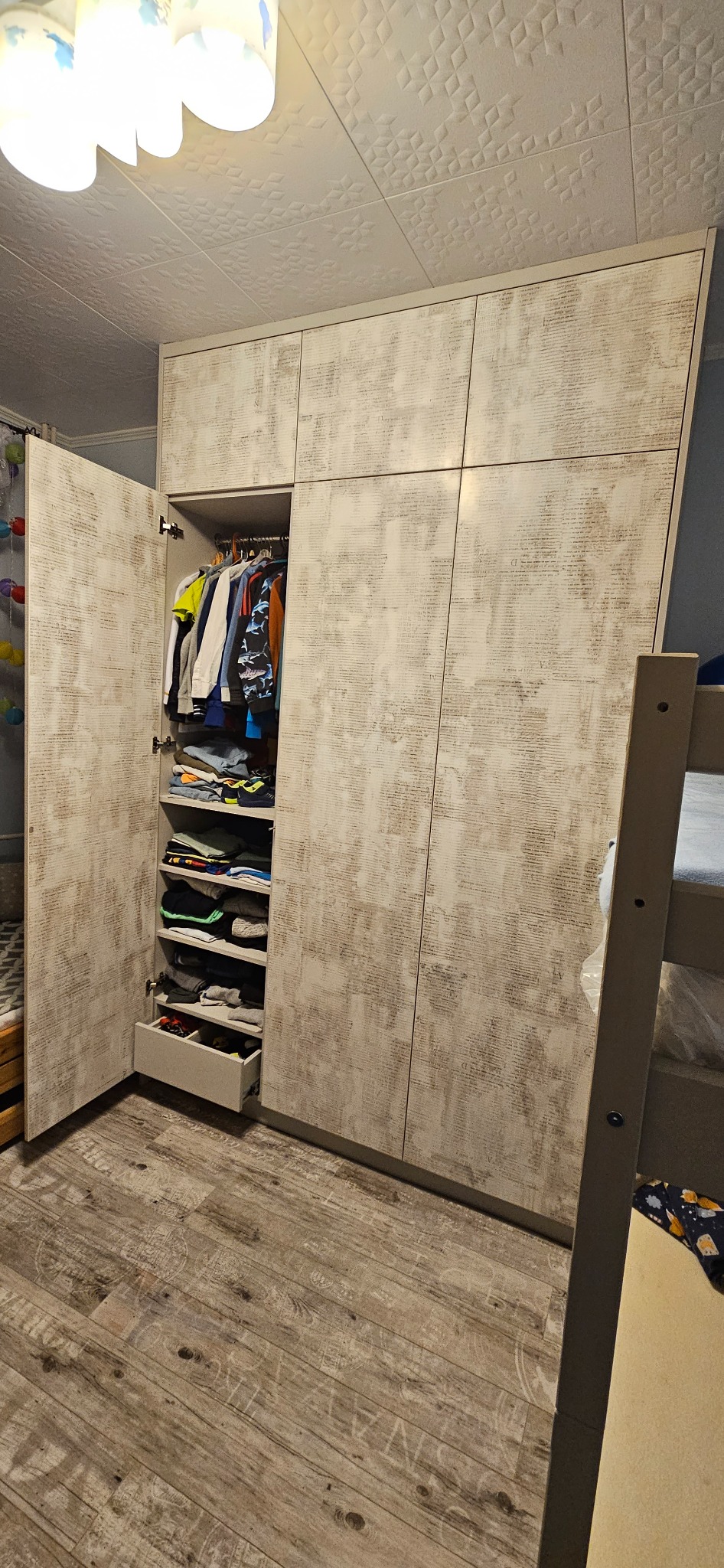 Wysoka szafa z otwieranymi drzwiami, wykończona w odcieniach beżu z delikatnym wzorem, wnętrze z półkami, szufladą i drążkiem na ubrania, widoczne ubrania i akcesoria, w tle łóżko piętrowe.