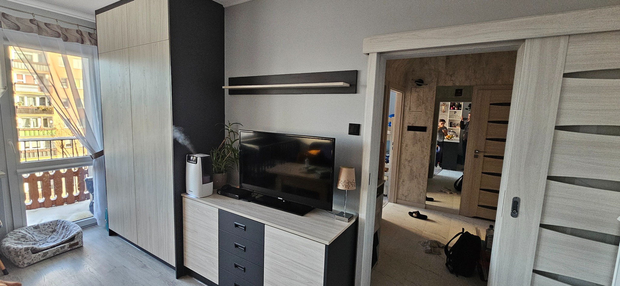 Nowoczesny pokój dzienny z szafą w zabudowie w kolorze jasnego drewna, obok komoda z szufladami w kolorze jasnego drewna i ciemnego grafitu, na niej telewizor, nawilżacz powietrza i roślina...