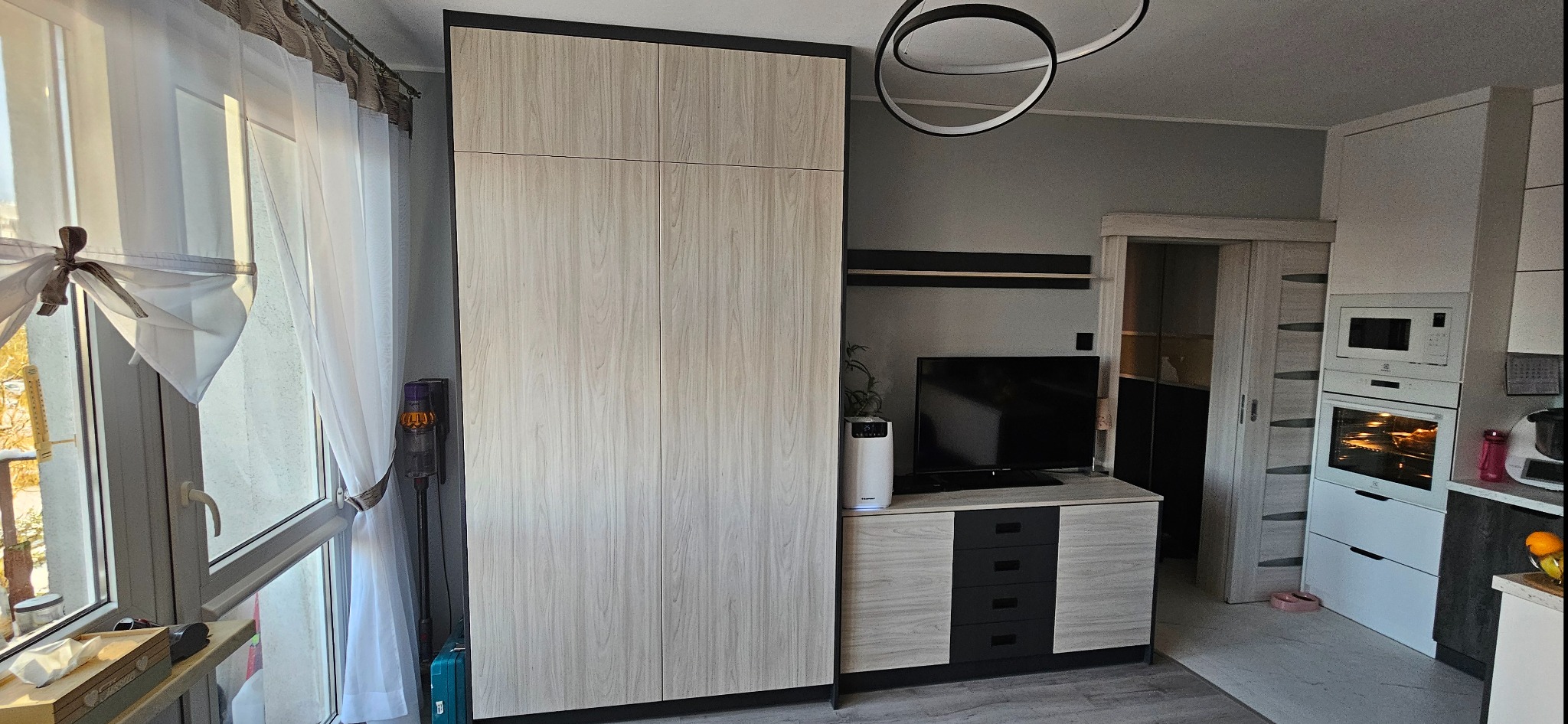 Jasna szafa na wymiar z czarnymi obramowaniami, dopasowana do szafki RTV w salonie z elementami kuchni w tle, oświetlona nowoczesną lampą sufitową.