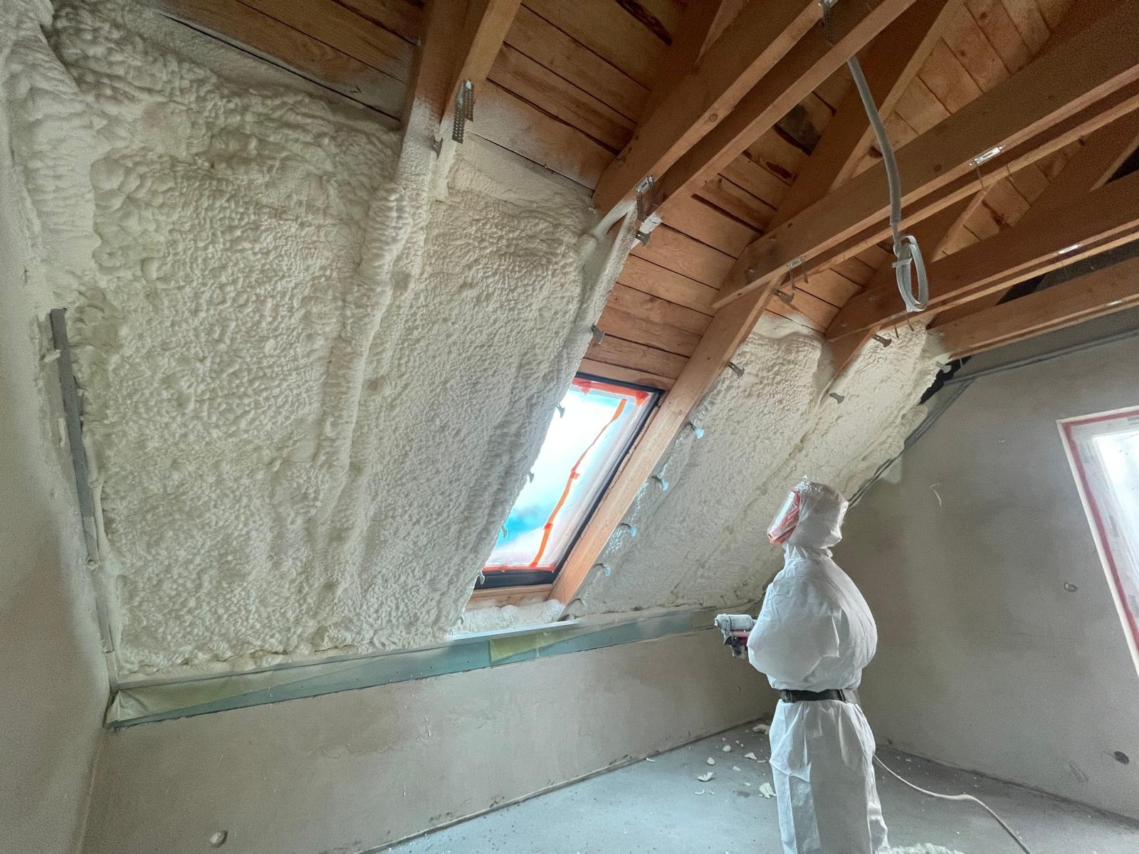 Poddasze po ociepleniu pianką PUR, widoczne drewniane elementy konstrukcyjne i okno dachowe. Pracownik w kombinezonie ochronnym z pistoletem do natrysku pianki.