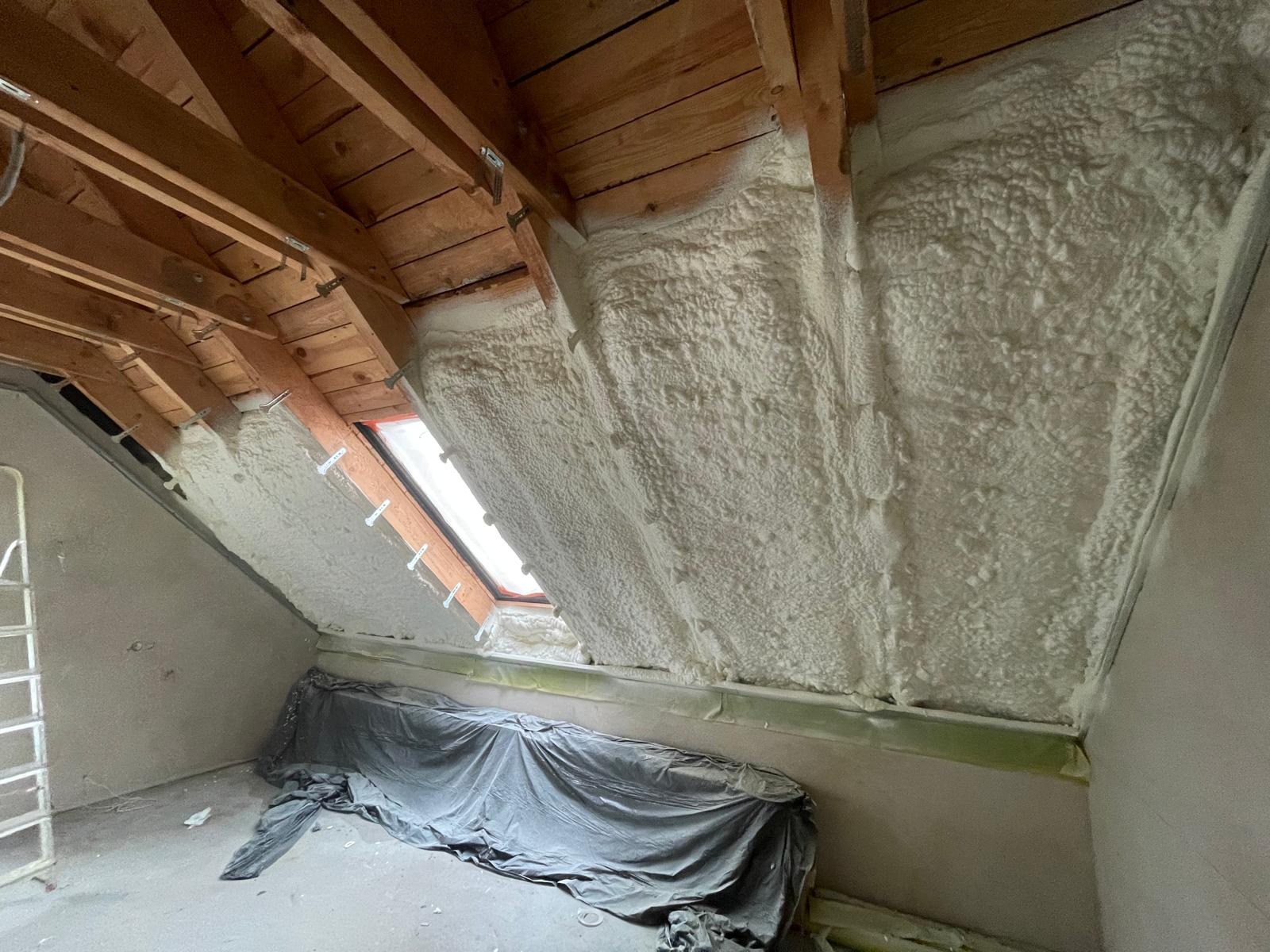 Poddasze z oknem połaciowym po natrysku pianki PUR, widoczne drewniane elementy konstrukcji dachu i zabezpieczona folią podłoga. Świeżo wykonana izolacja.