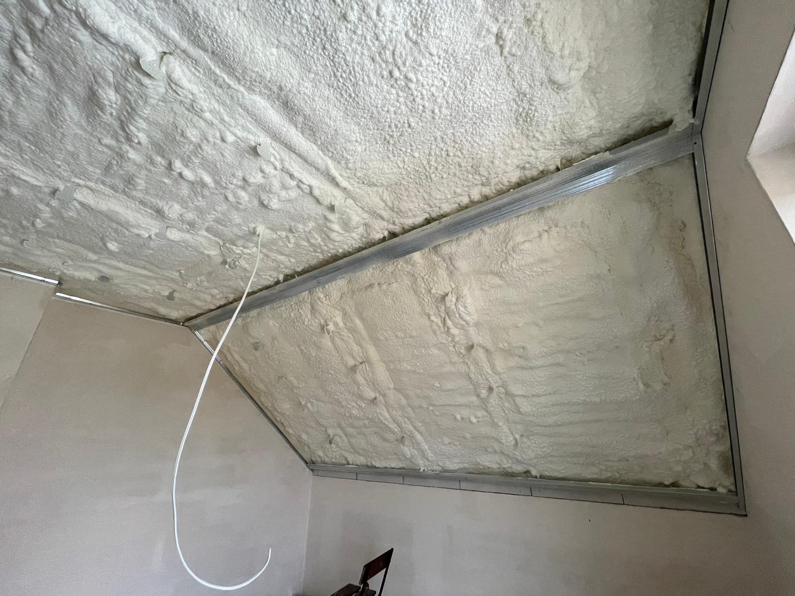 Poddasze po ociepleniu pianką PUR, widoczne metalowe profile konstrukcyjne. Biała, nierówna powierzchnia pianki pokrywa skośny sufit. W rogu pomieszczenia stoi krzesło.