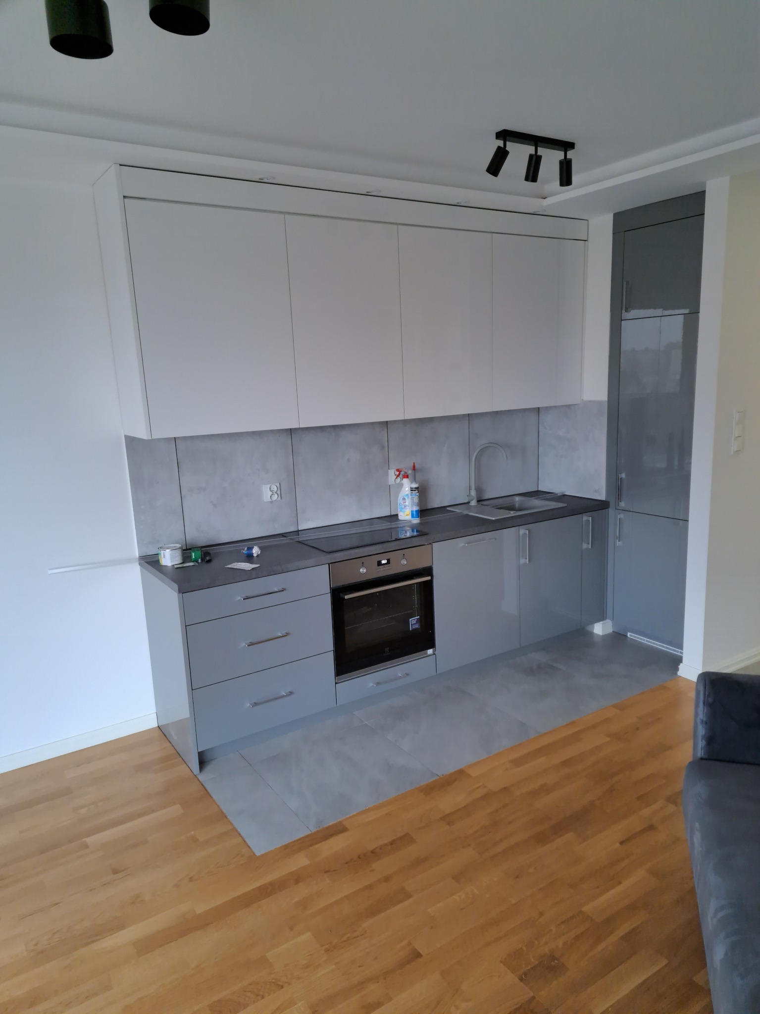 Nowoczesna kuchnia w odcieniach szarości i bieli z drewnianą podłogą; widoczne szafki górne i dolne, płyta indukcyjna, piekarnik oraz akcesoria na blacie.