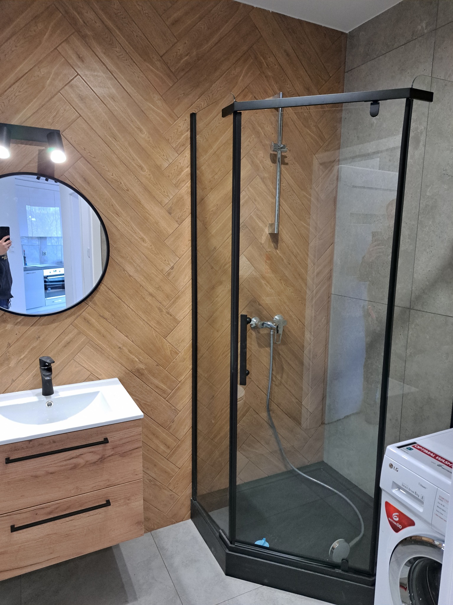 Nowoczesna łazienka z kabiną prysznicową z czarnymi profilami, umywalką z szafką z drewnopodobnym frontem ułożonym w jodełkę, okrągłym lustrem z oświetleniem i pralką LG w rogu