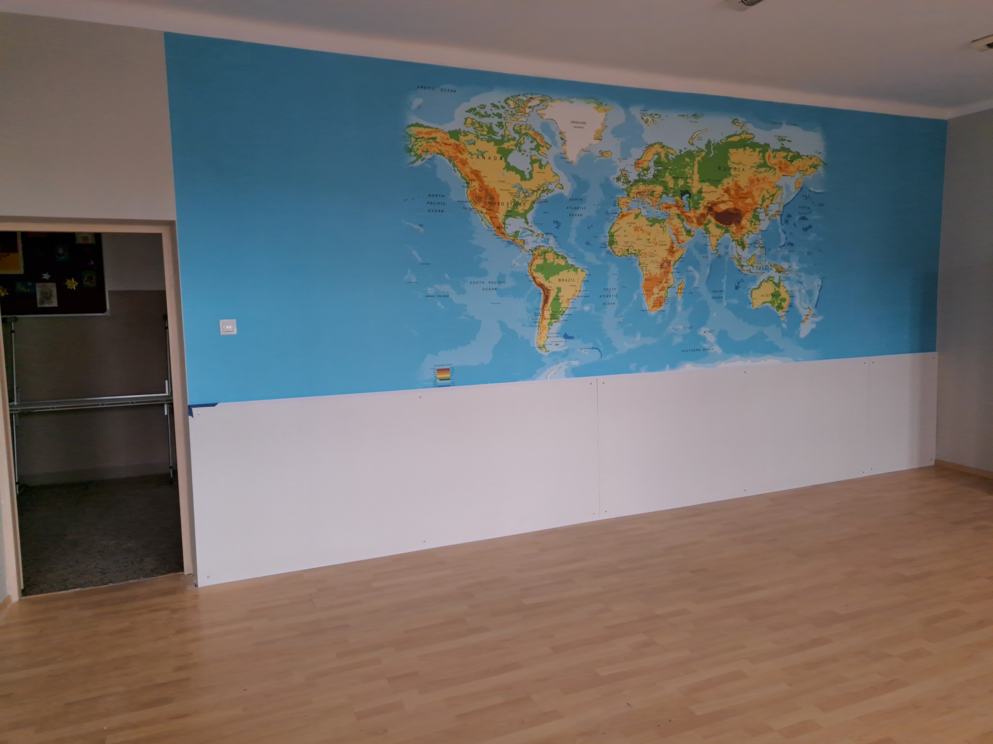 Ściana w pokoju z dużą mapą świata, dolna część ściany wykończona białymi panelami, widoczna podłoga z paneli i fragment otwartych drzwi do pomieszczenia gospodarczego.