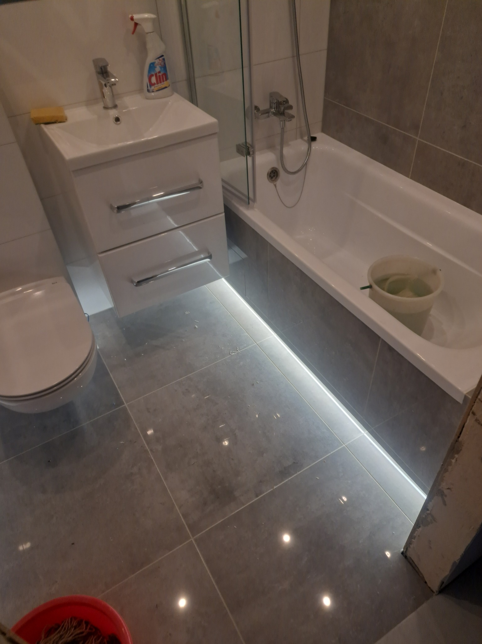 Nowoczesna łazienka z podświetleniem LED pod wanną i szafką umywalkową, szare płytki podłogowe, biała armatura i akcesoria.