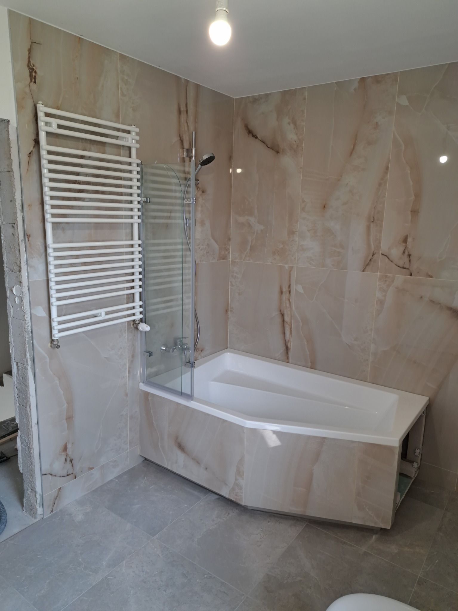 Wykończona łazienka z beżowymi płytkami imitującymi marmur, białą narożną wanną z parawanem nawannowym oraz grzejnikiem drabinkowym na ścianie, widoczne szare płytki podłogowe.