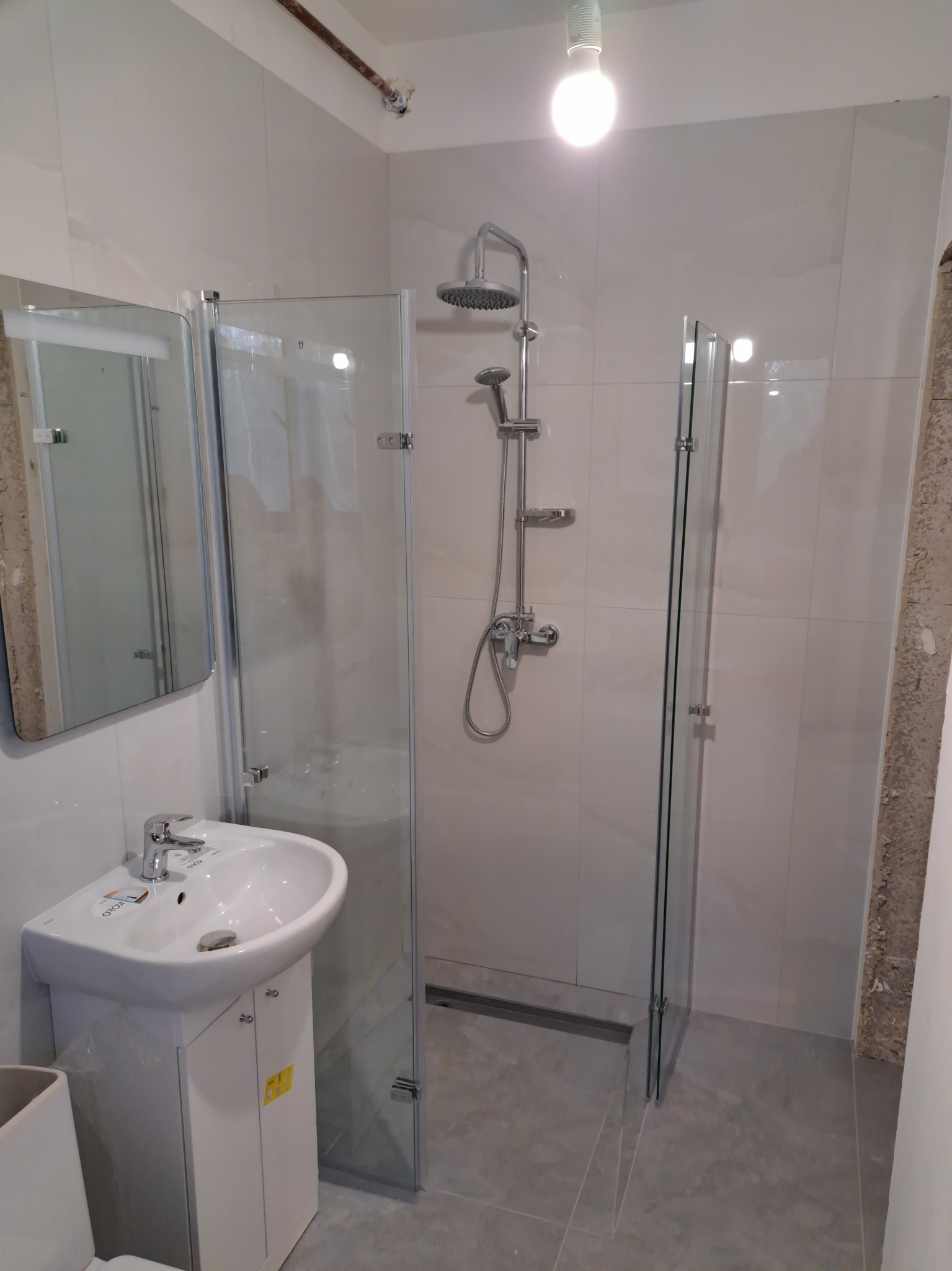 Nowoczesna łazienka z kabiną prysznicową typu walk-in, umywalką na szafce i szarymi płytkami podłogowymi, widoczna rura pod sufitem.