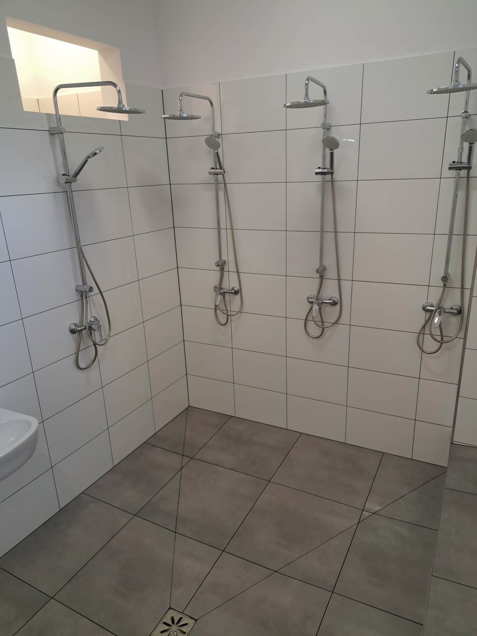 Wnętrze publicznej łazienki z czterema kabinami prysznicowymi z chromowanymi bateriami i deszczownicami, wykończone białymi płytkami ściennymi i szarymi płytkami podłogowymi z odpływem liniowym.