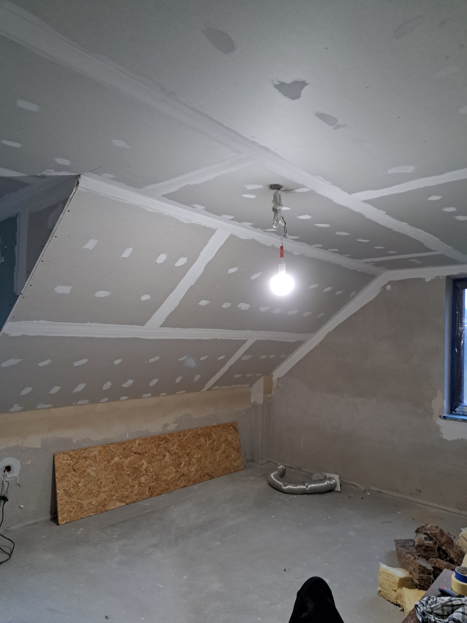 Poddasze w trakcie wykańczania: płyty gipsowo-kartonowe na skosach i suficie, widoczne łączenia wzmocnione taśmą, surowy beton na ścianach, wisząca żarówka, fragment okna, płyta OSB leżąca...