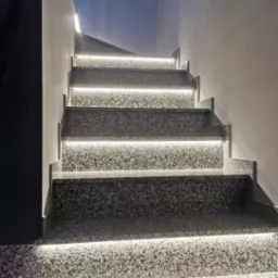 Granitowe schody z subtelnym, białym oświetleniem LED wzdłuż krawędzi stopni, oświetlające ścianę i tworzące nowoczesny efekt wizualny.