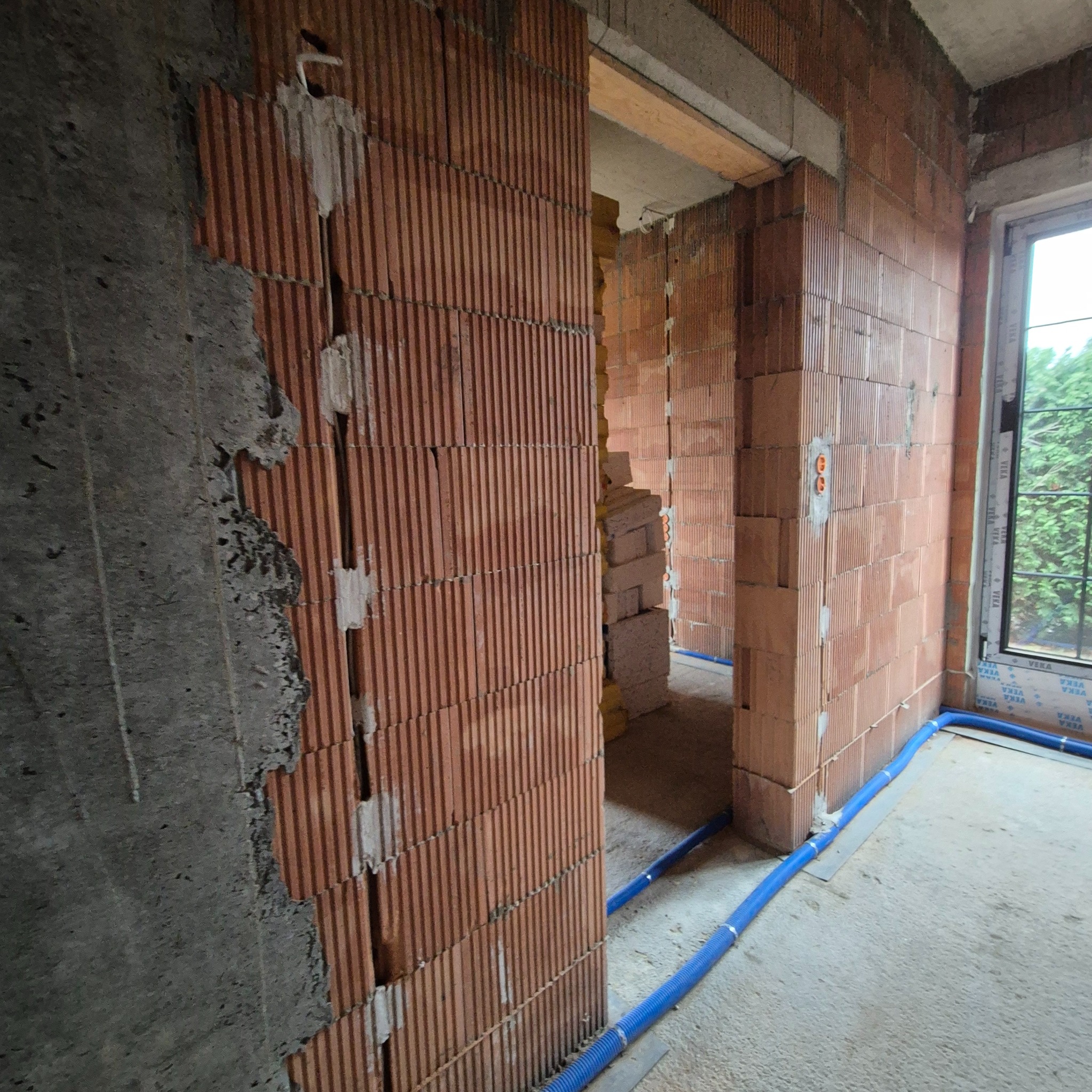 Wnętrze w trakcie budowy z ceglanymi ścianami, widoczne pionowe bruzdy z przewodami elektrycznymi zamaskowanymi gipsem, niebieskie rury instalacyjne na podłodze i okno z widokiem na zieleń.