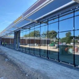 Montaż wielkoformatowych okien aluminiowych na elewacji budynku, widoczni pracownicy w odblaskowych kamizelkach, teren budowy.