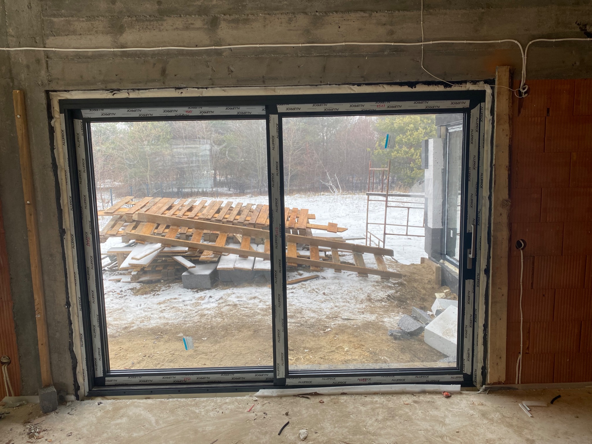 Montaż przesuwnych okien aluminiowych z czarnymi ramami w surowym stanie, widok na zewnątrz z paletami i śniegiem.