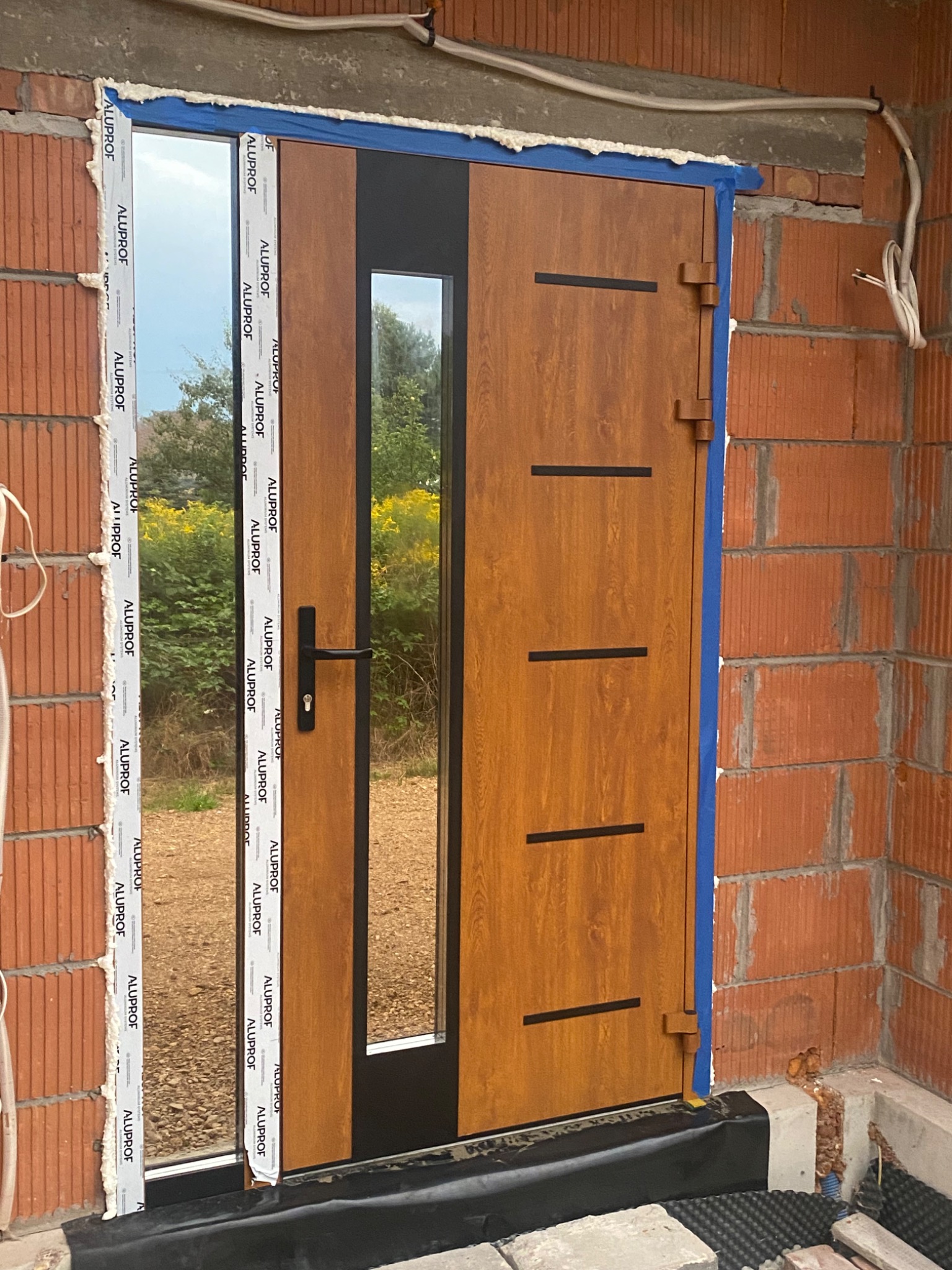 Montaż drzwi zewnętrznych z przeszkleniem, widoczne pianka montażowa, taśma malarska i ceglany mur w surowym stanie, elementy wykończeniowe Aluplast.