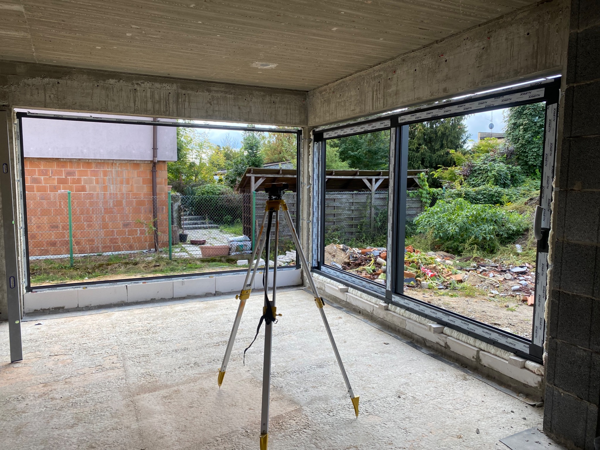 Surowy stan wnętrza z trzema oknami aluminiowymi z czarnymi ramami, widok na zewnątrz z zielenią, na pierwszym planie niwelator na statywie, betonowe ściany i sufit w stanie deweloperskim.