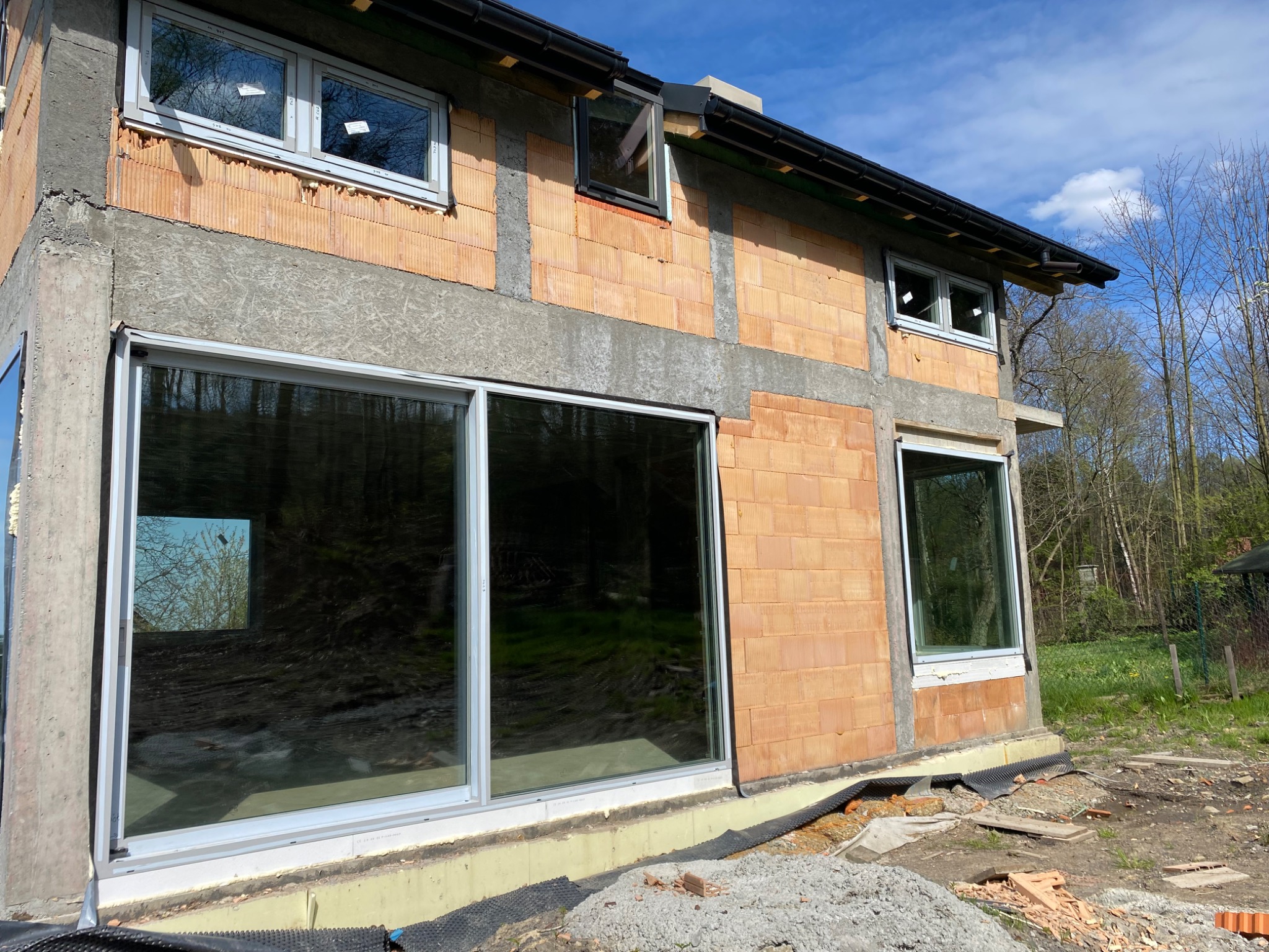 Montaż dużych okien przesuwnych w surowym budynku z czerwonej cegły, widoczne szare ramy okienne i betonowe wzmocnienia, teren wokół budynku przygotowany pod dalsze prace wykończeniowe.