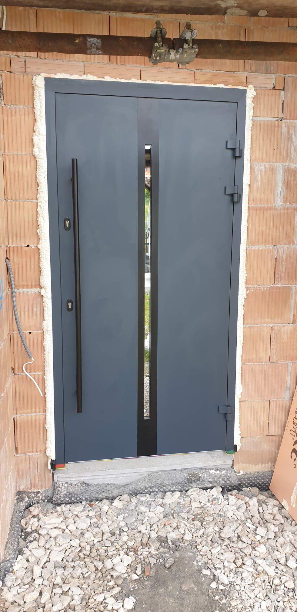 Szare, dwuskrzydłowe drzwi z długim, pionowym przeszkleniem pośrodku, zamontowane w surowym murze z czerwonej cegły, z widoczną pianką montażową i gruzem u podstawy.