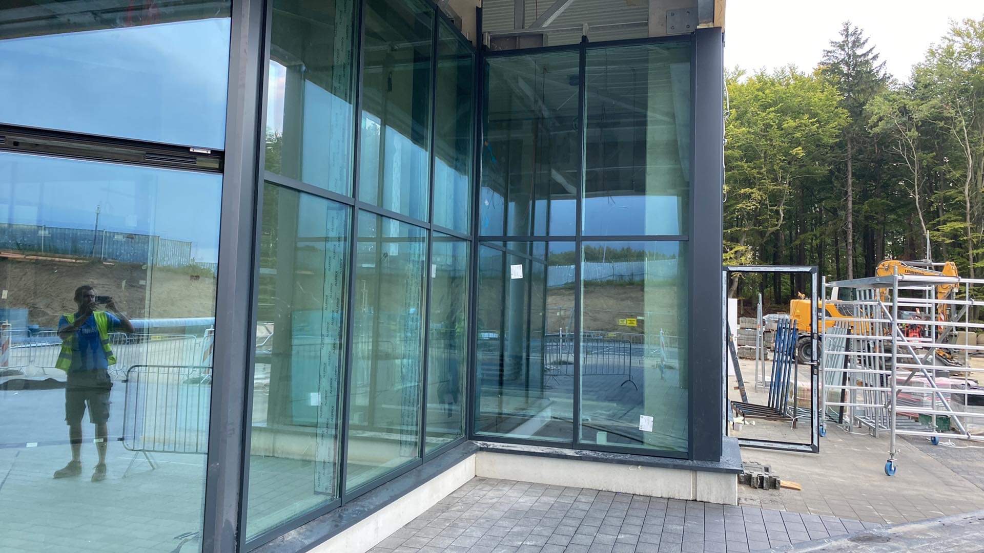 Narożne, duże przeszklenie budynku w czarnych ramach, widoczne rusztowania i prace budowlane w tle, odbicie osoby robiącej zdjęcie w szybie.