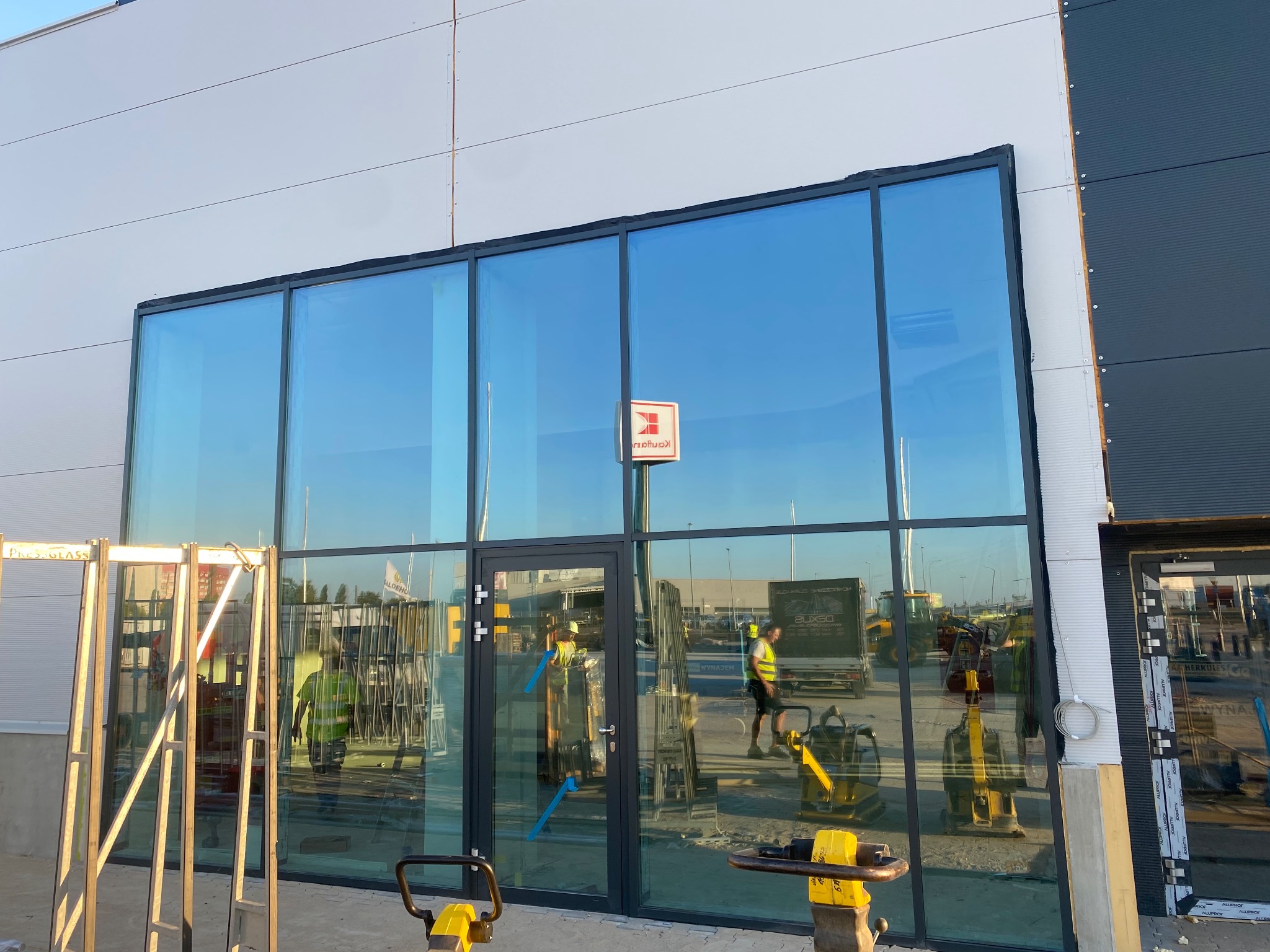 Montaż dużych, szklanych okien w budynku przemysłowym; widoczne rusztowania, pracownicy w kamizelkach odblaskowych i sprzęt budowlany.