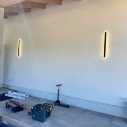 Dwa pionowe, podłużne kinkiety LED, zamontowane na jasnej ścianie zewnętrznej pod drewnianą konstrukcją z belek stropowych. Na podłodze leżą skrzynki narzędziowe, torba i hulajnoga.