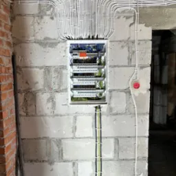 Rozdzielnica elektryczna z licznymi przewodami ułożonymi w peszlach i przymocowanymi do surowej ściany z betonu komórkowego, widoczne także gniazda elektryczne z czerwonymi zaślepkami.