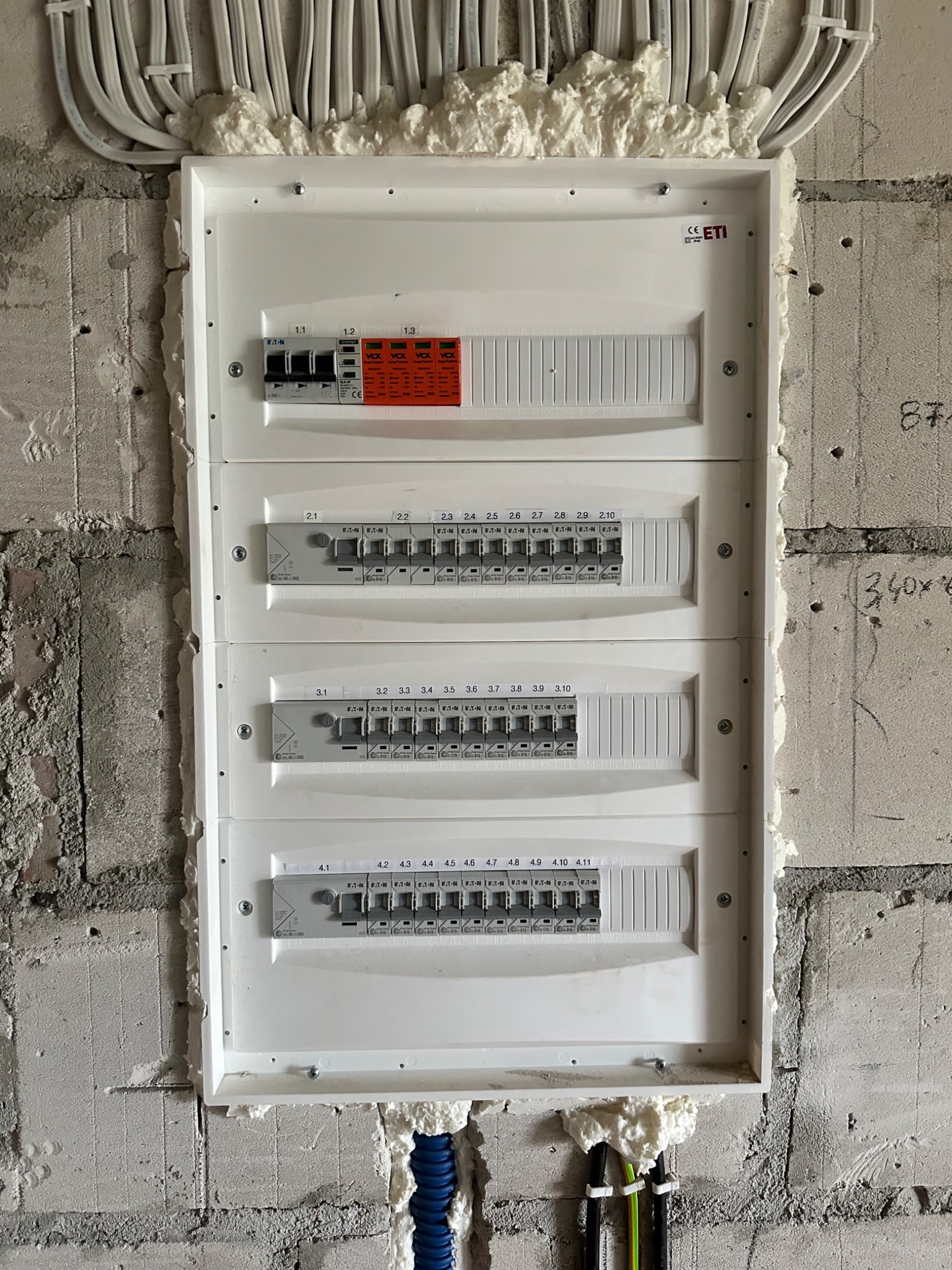 Rozdzielnica elektryczna z zabezpieczeniami, zamontowana na surowej ścianie, zabezpieczona pianką montażową, widoczne kable w peszlach