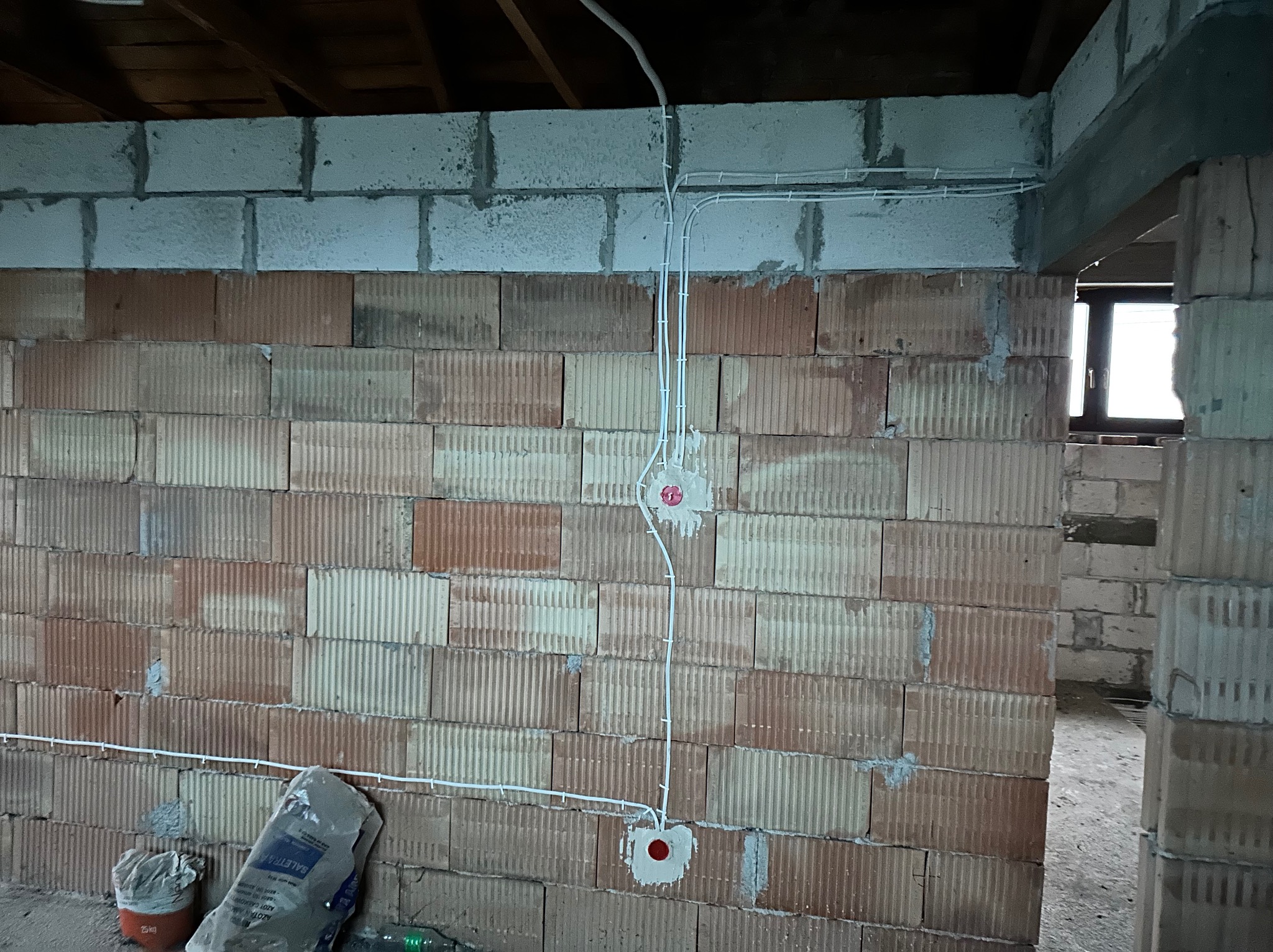 Ściana z pustaków z widocznymi, częściowo zamontowanymi przewodami elektrycznymi w białych peszlach, zakończonymi puszkami instalacyjnymi z czerwonymi zaślepkami. Na podłodze worek z zaprawą.